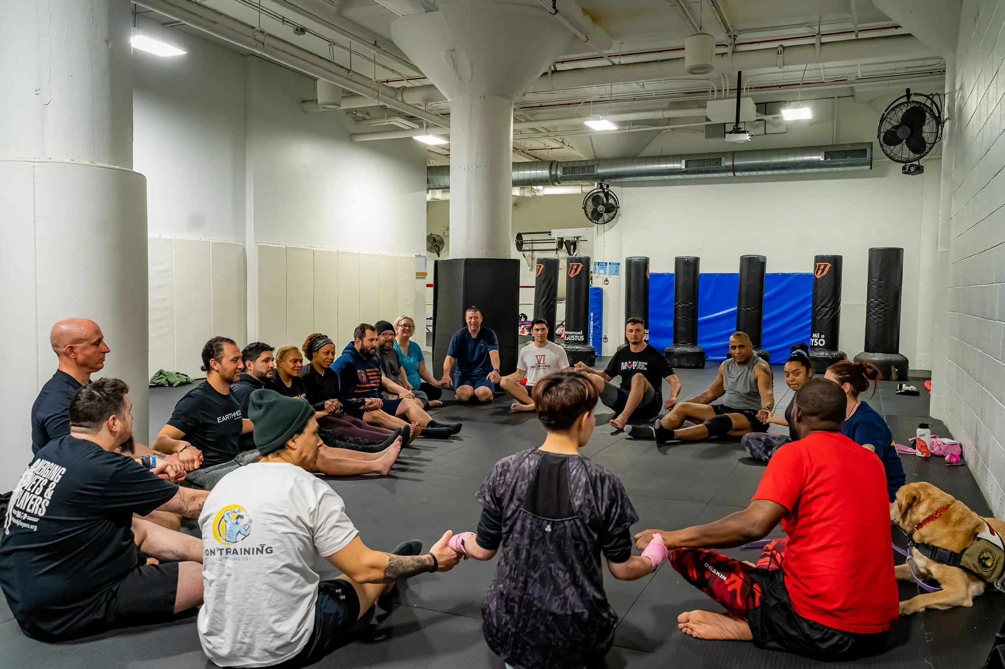Group of people sitting cross-legged in a circle inside a gym, holding hands during a session, with a dog lying beside a person in the foreground.