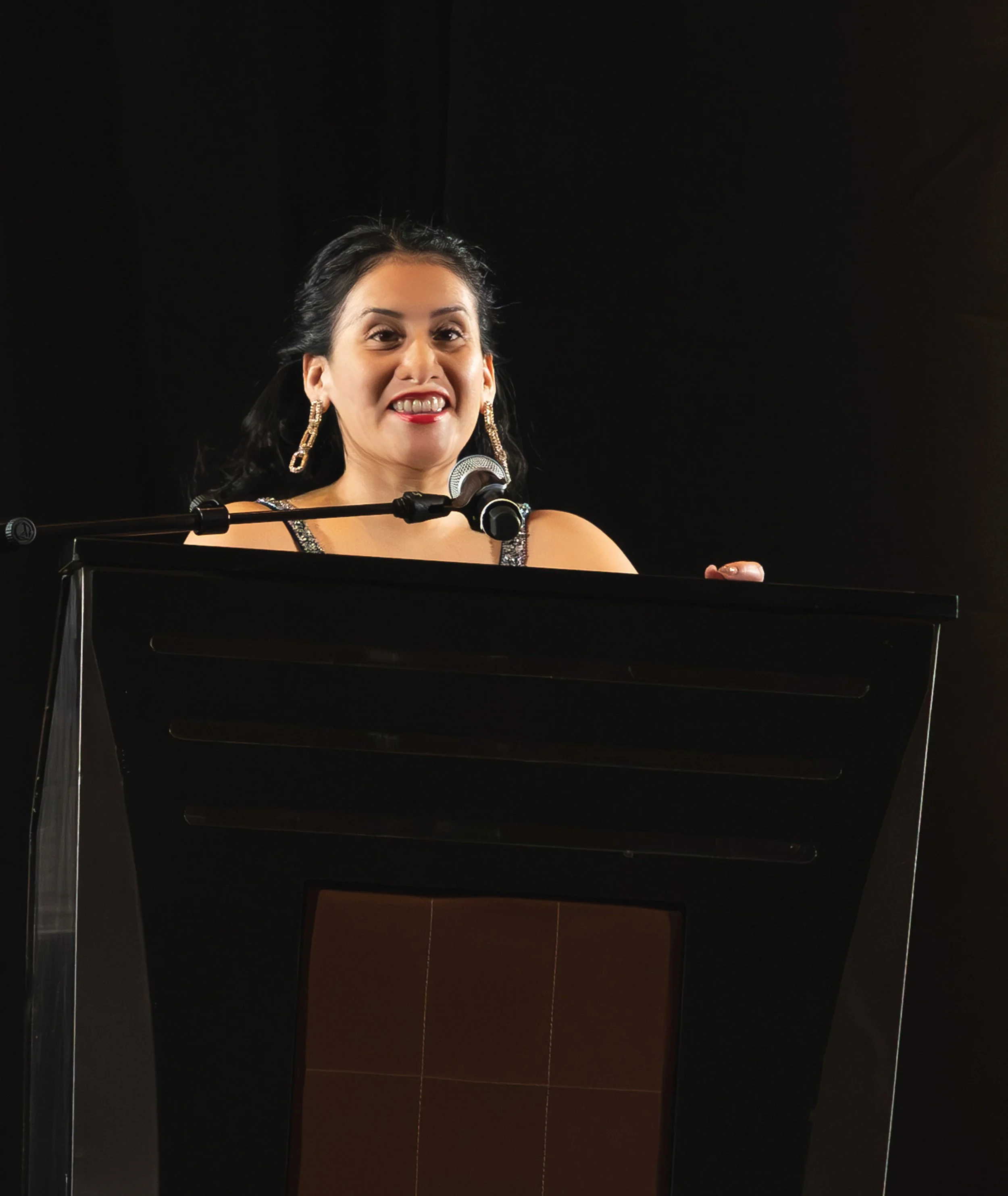 A woman speaking at a podium with a microphone, wearing a black dress and gold earrings, smiling.