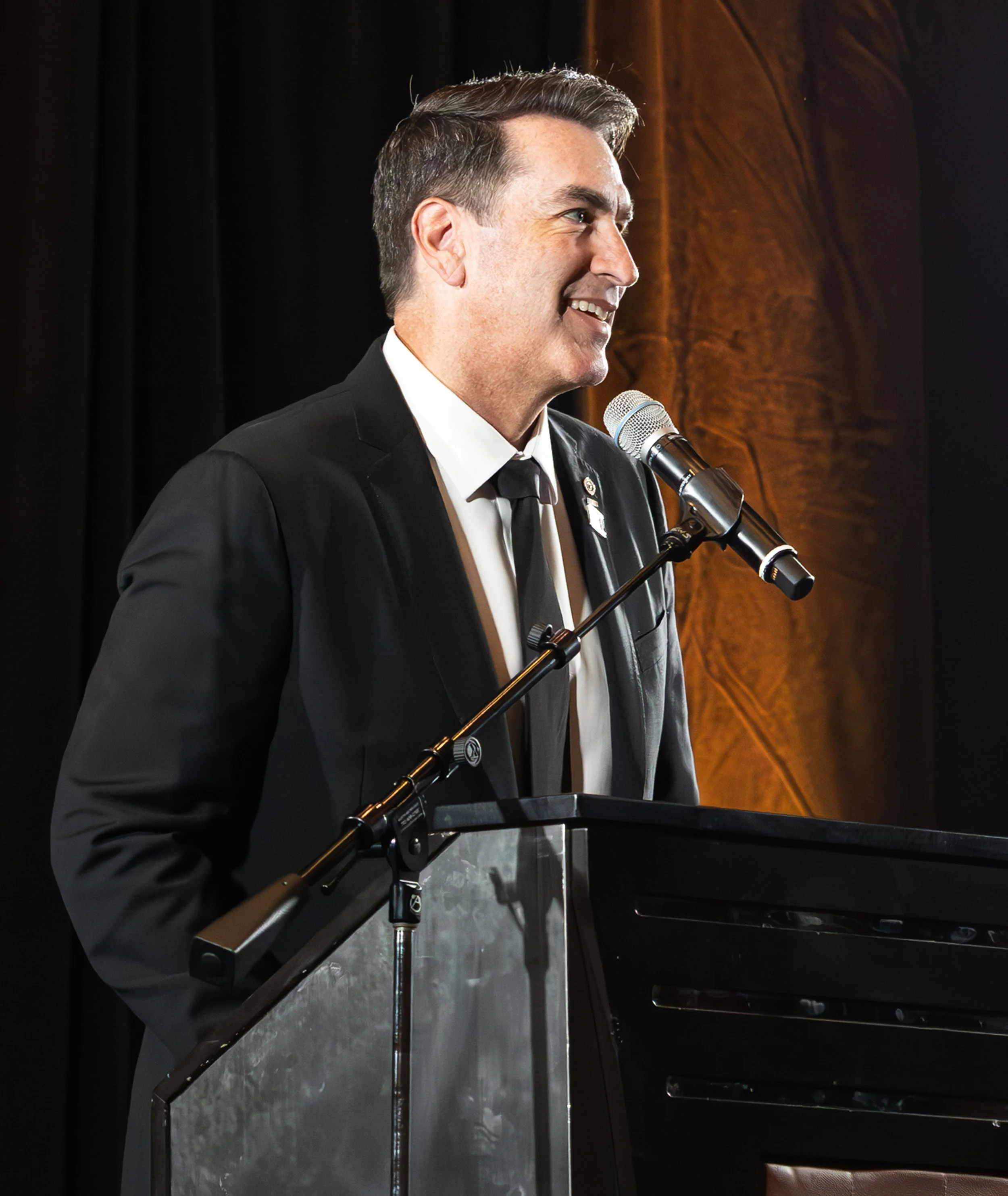 Man in a black suit and tie smiling while speaking at a podium with a microphone.