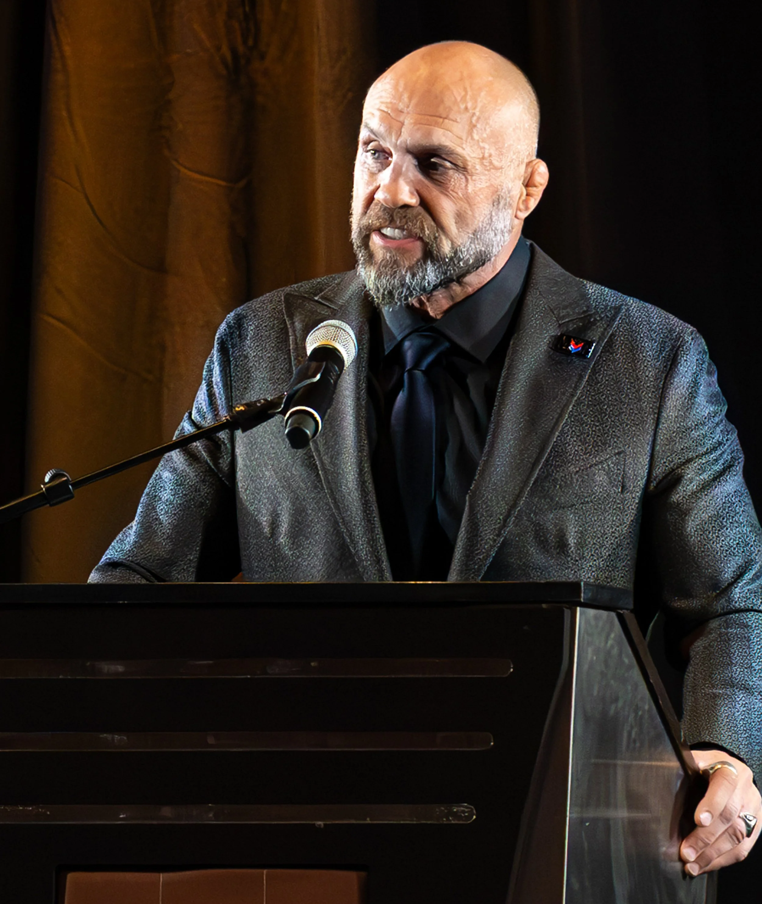 A man with a bald head and beard speaking at a podium with a microphone, wearing a dark suit and a tie.
