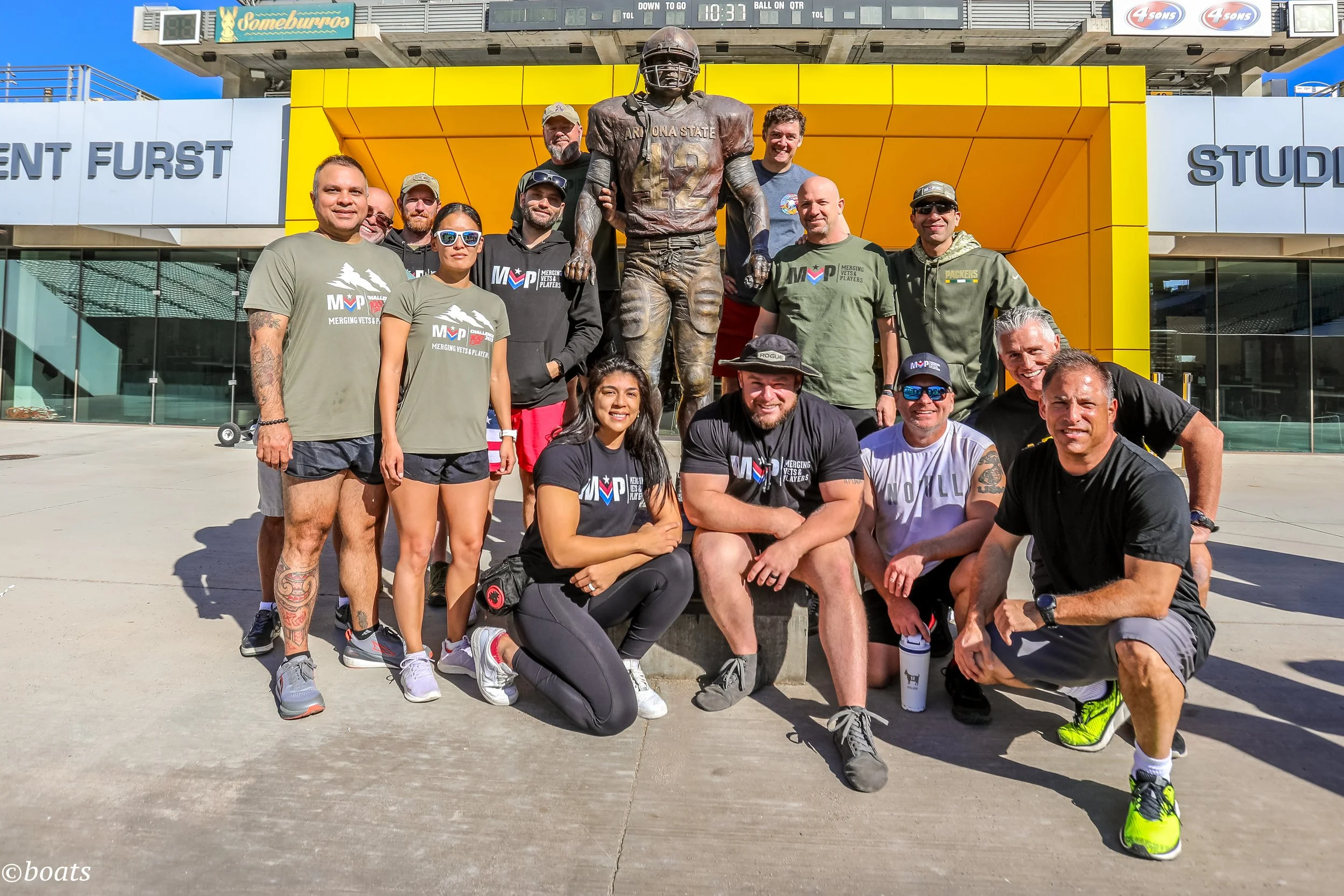 Group of people gathered outside a stadium with a large statue of a football player in front of a yellow and white building. The group appears to be participating in a sporting event or team activity, with some wearing matching shirts that say 'Merging Vets & Players.'