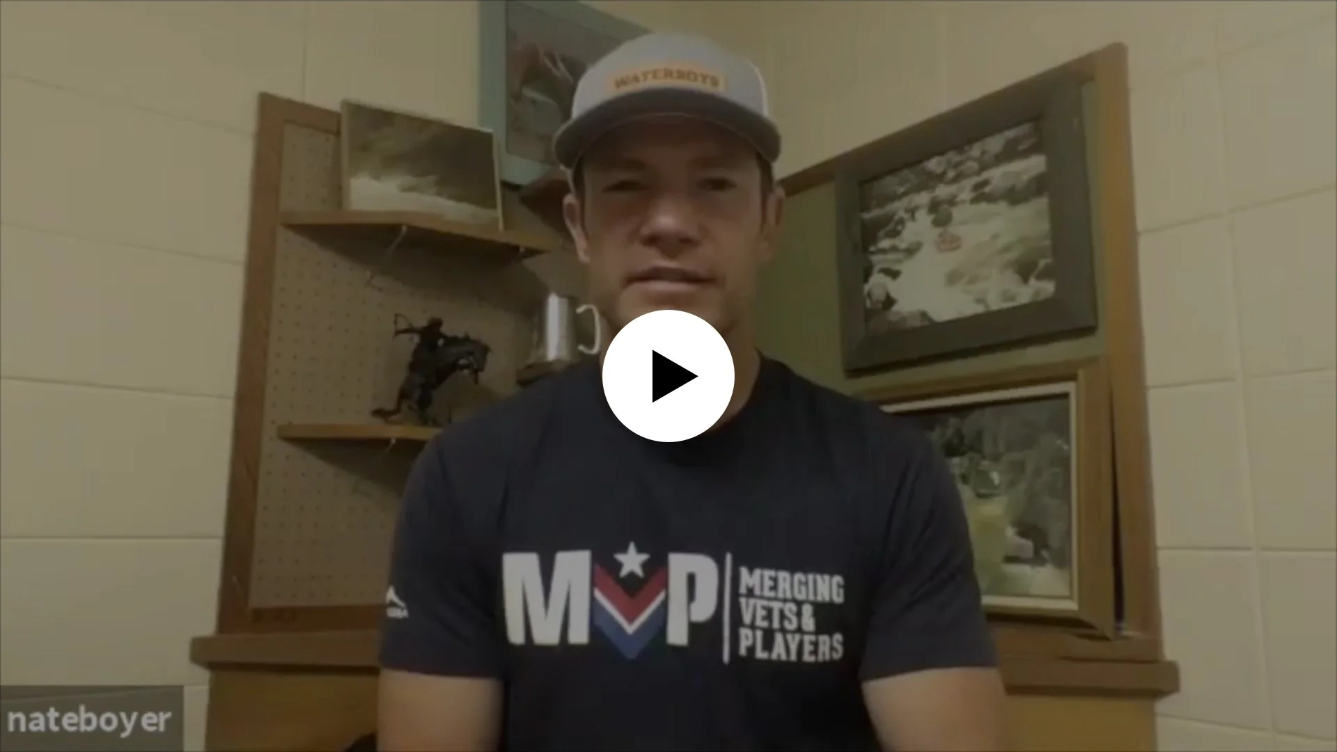 Young man wearing a baseball cap and a black T-shirt with 'MVP Merging Vets & Players' logo, sitting in a room with framed artwork and a wooden shelf behind him.
