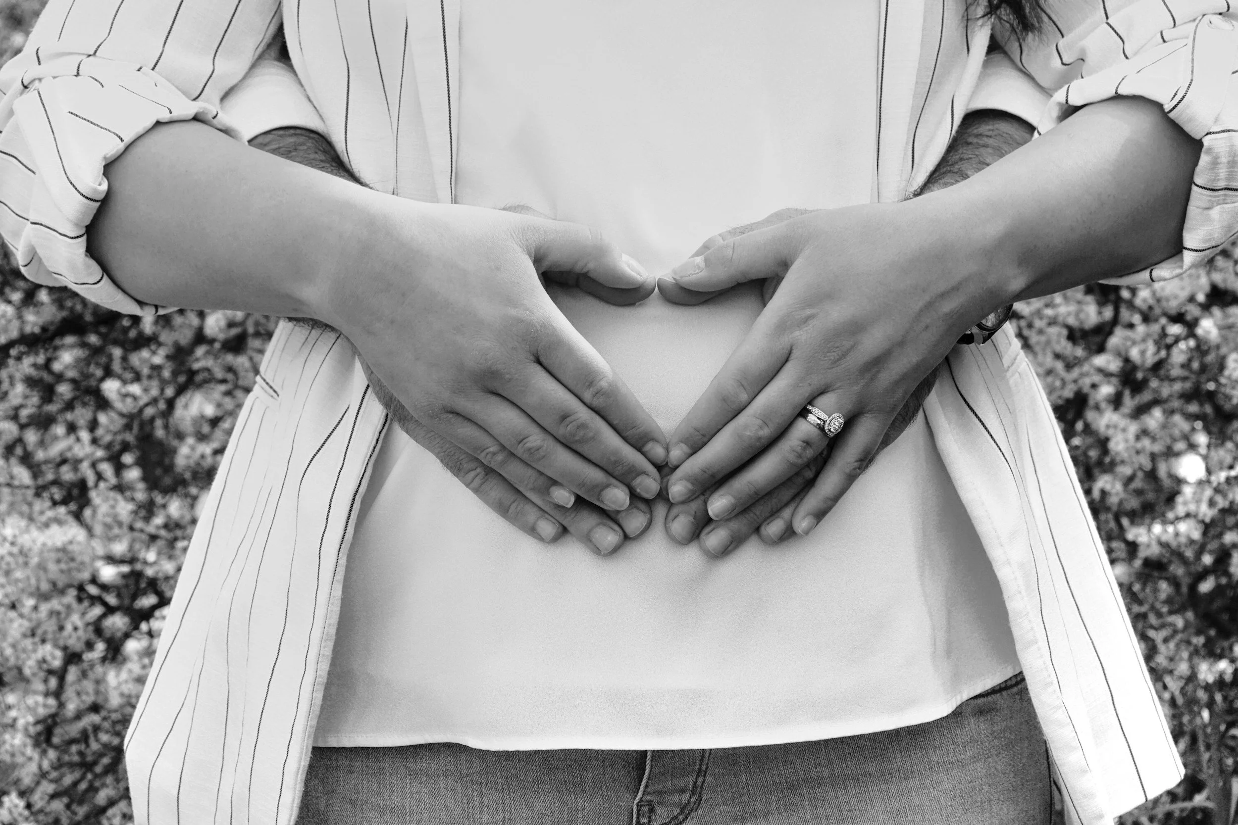 Close-up of a pregnant woman's belly with hands from a couple forming a heart shape around her baby bump. The woman is wearing a ring on her left ring finger. The photo is in black and white with a natural outdoor background.