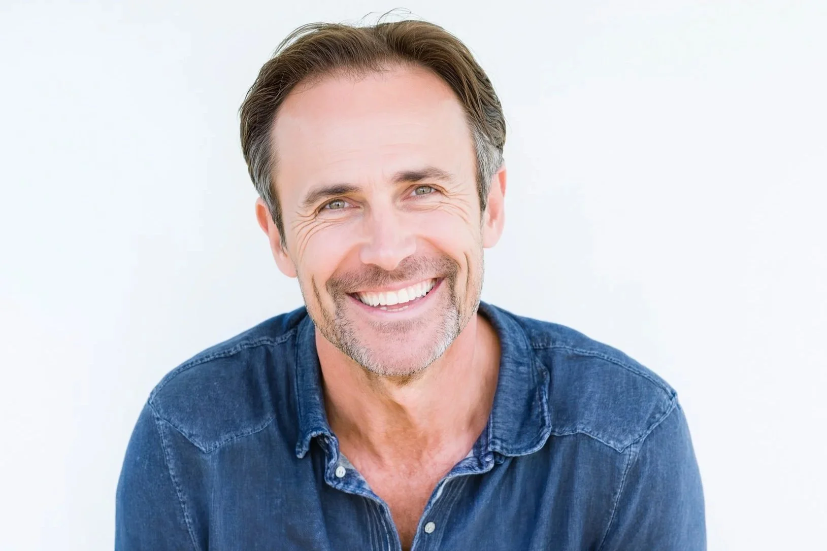 Smiling middle-aged man with dark hair and stubble, wearing a blue denim shirt, against a plain white background.