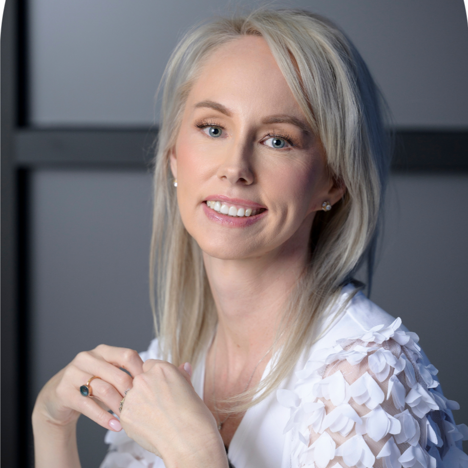 Close-up of a woman with blonde hair, smiling and wearing a white top with textured, petal-like fabric on the shoulders, sitting against a dark background.