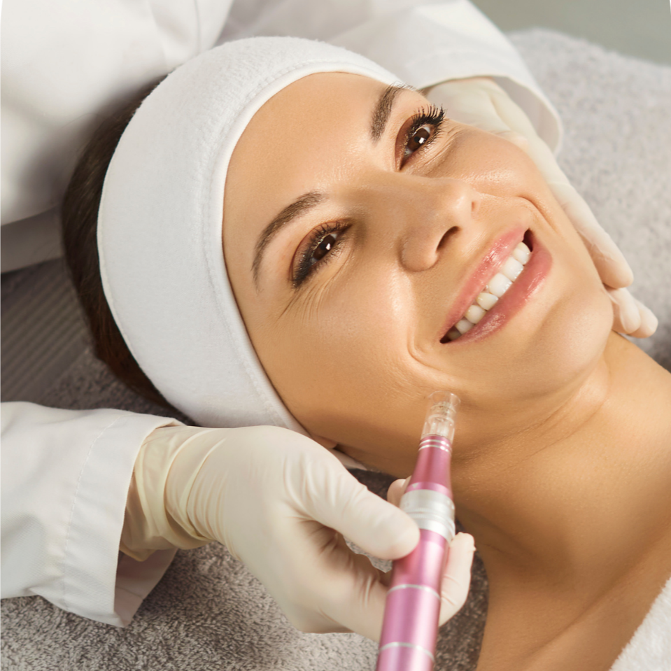 A woman receiving a facial treatment, lying on a bed with her head resting on a pillow, smiling as a skincare professional applies a facial massage or treatment with a pink tool.