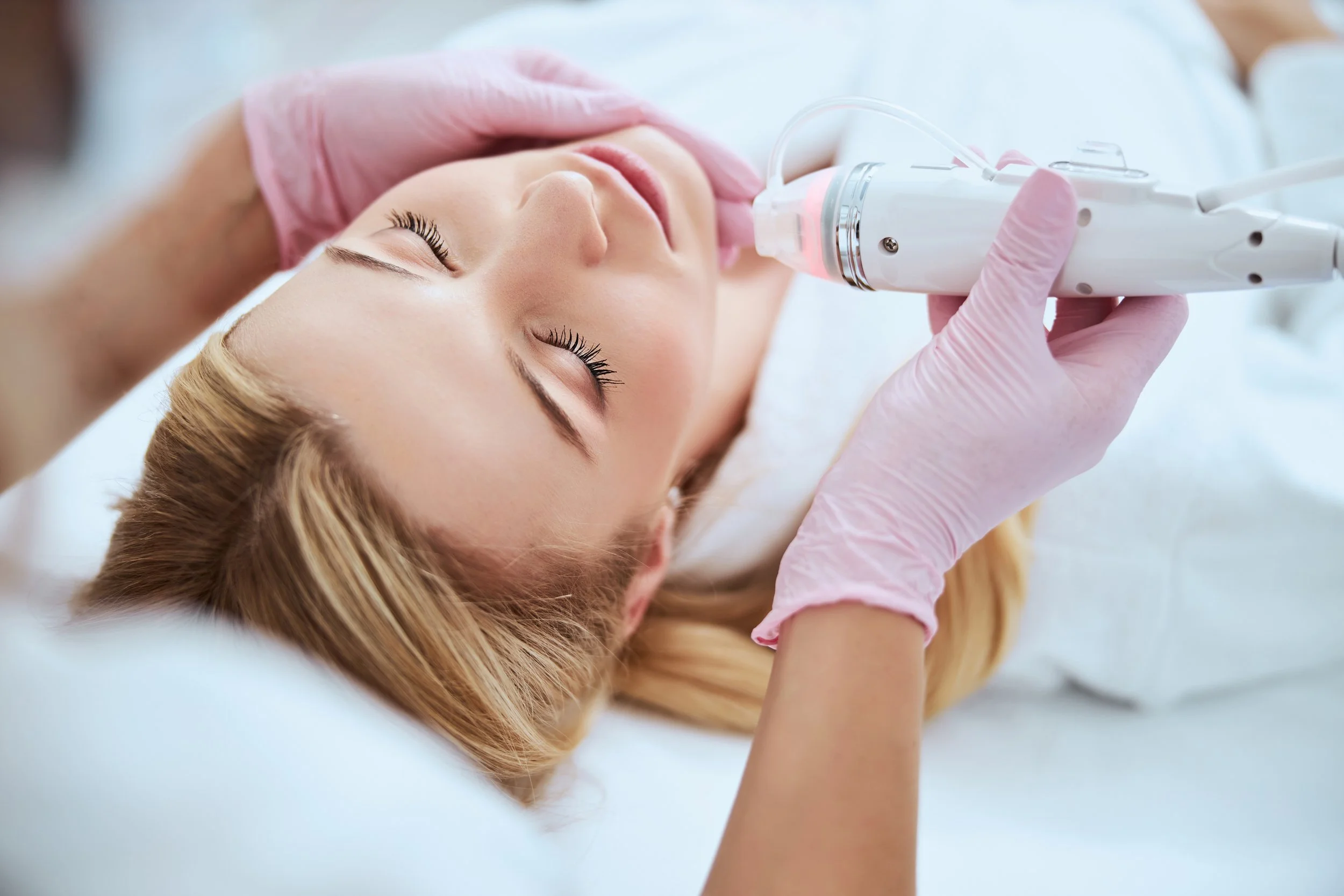 A woman lying down with her eyes closed receiving a facial treatment with a handheld device from a practitioner wearing pink gloves.