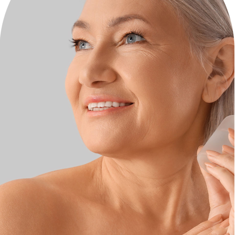 Close-up of a smiling older woman with gray hair, blue eyes, and fair skin, holding a white object near her neck.