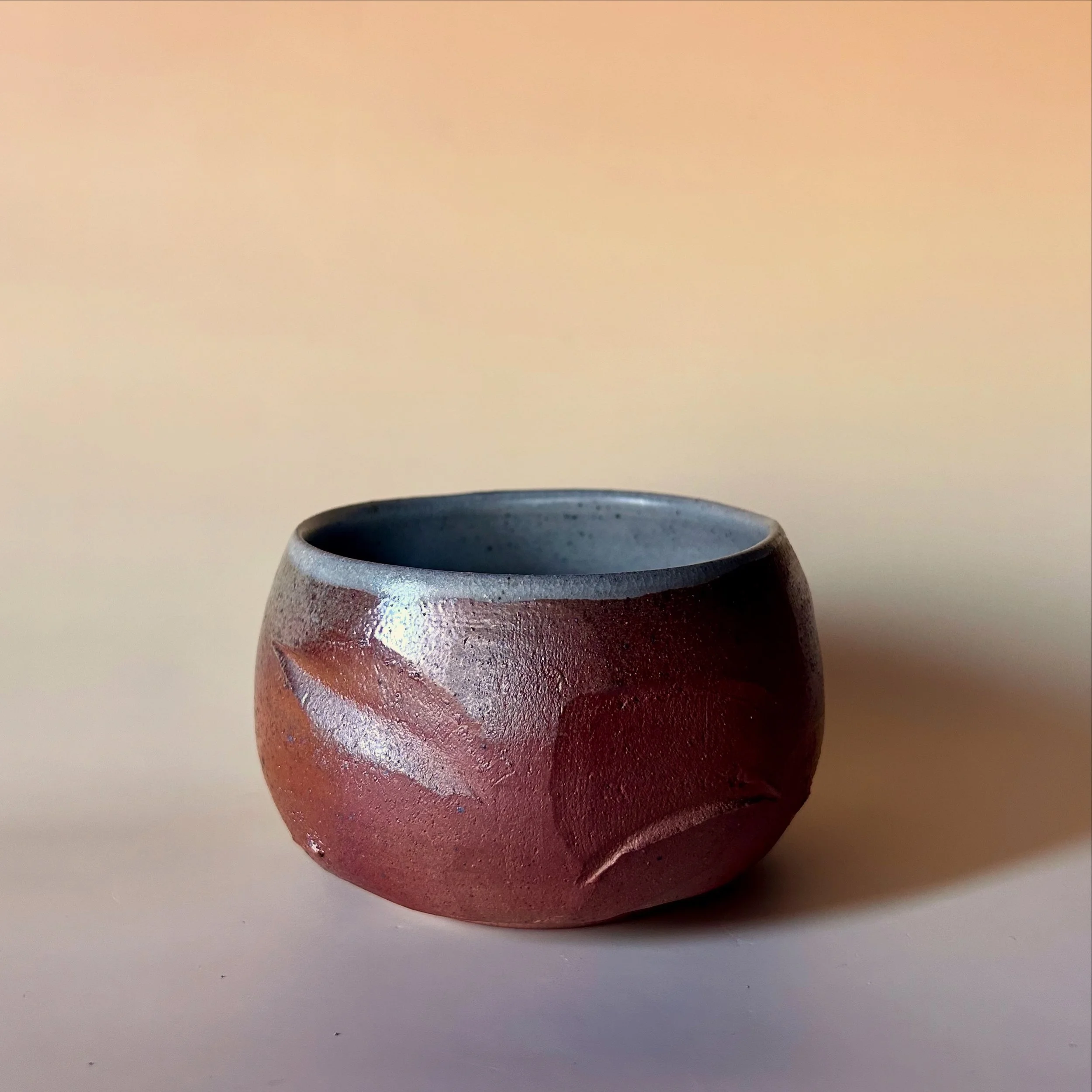 A small round wood fired ceramic tea bowl with a textured reddish-brown exterior and a ice celadon  interior, placed on a plain surface with a light-colored background.