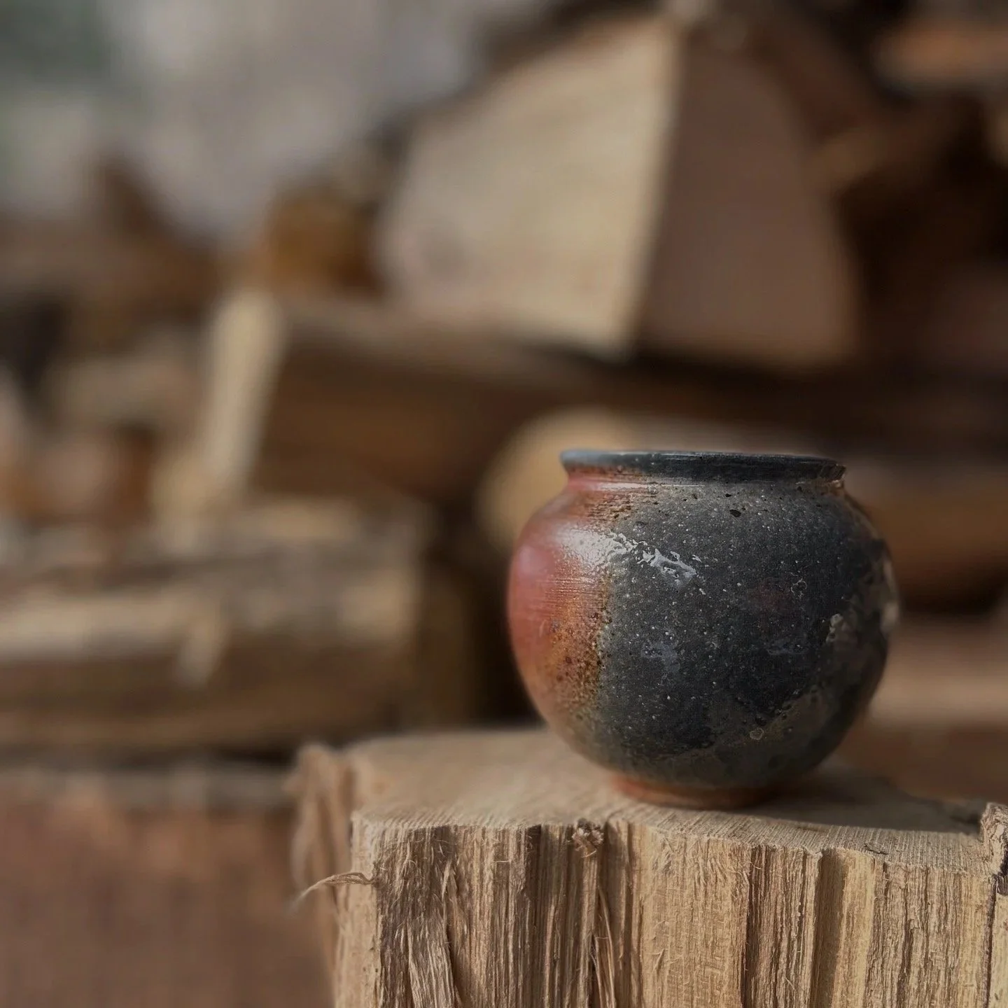 A mini soda-fired moon jar with blue, grey, and orange coloring,  placed on a piece of wood with a blurred background of stacked wood logs.