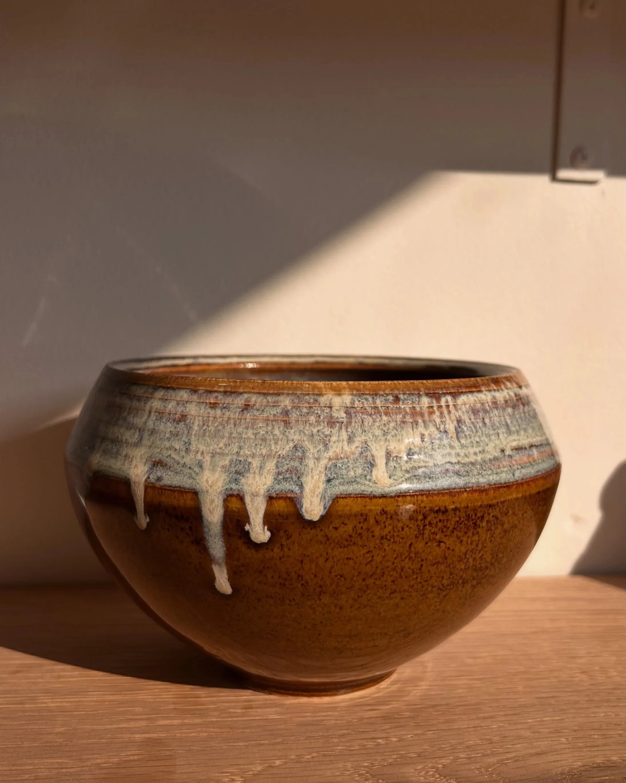 An electric-fired ceramic carinated bowl with a brown glaze and a decorative glaze drip pattern, placed on a wooden surface with sunlight casting shadows.