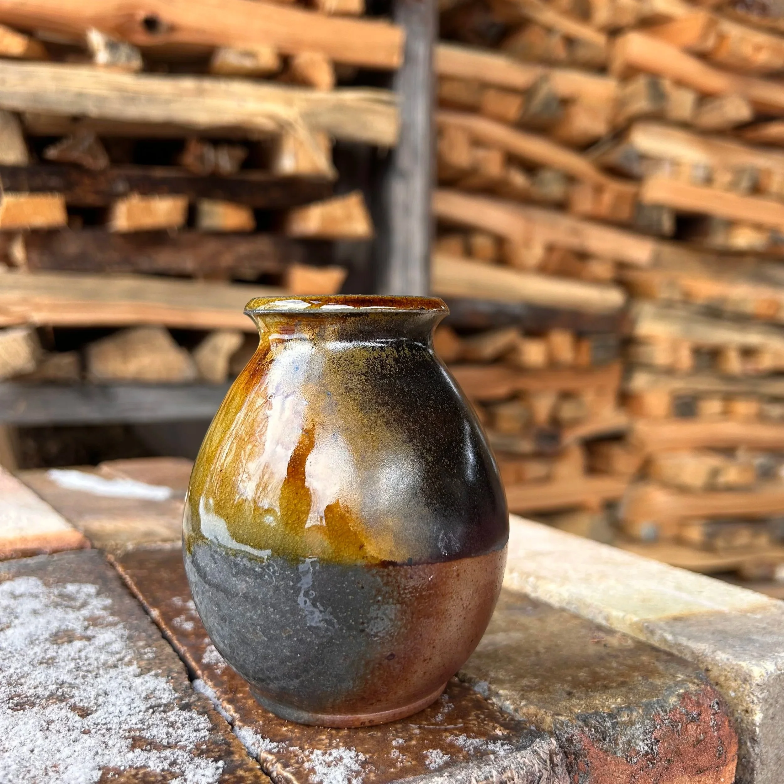 Colorful ceramic wood fired bud vase with dark and earthy tones from tenmoku glaze, sitting on a rustic brick surface with a background of stacked firewood.