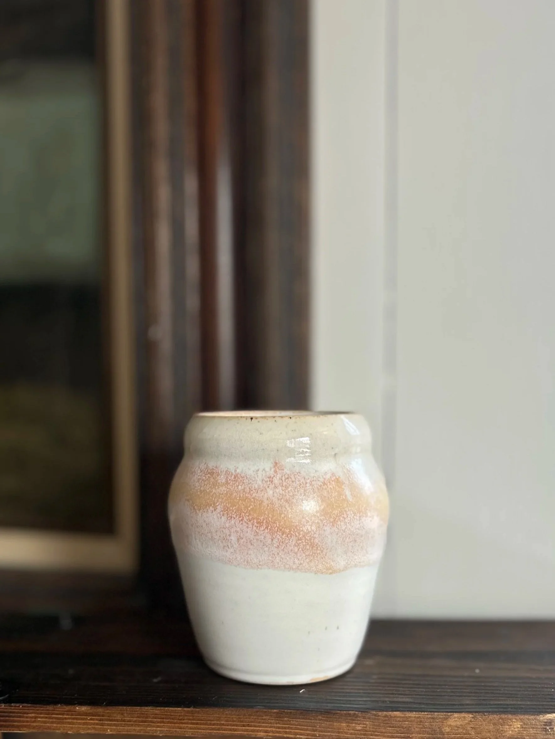 A small electric-fired ceramic vase, glazed in stone white with a band of warm orange creme glaze near the top, placed on a dark wooden surface.