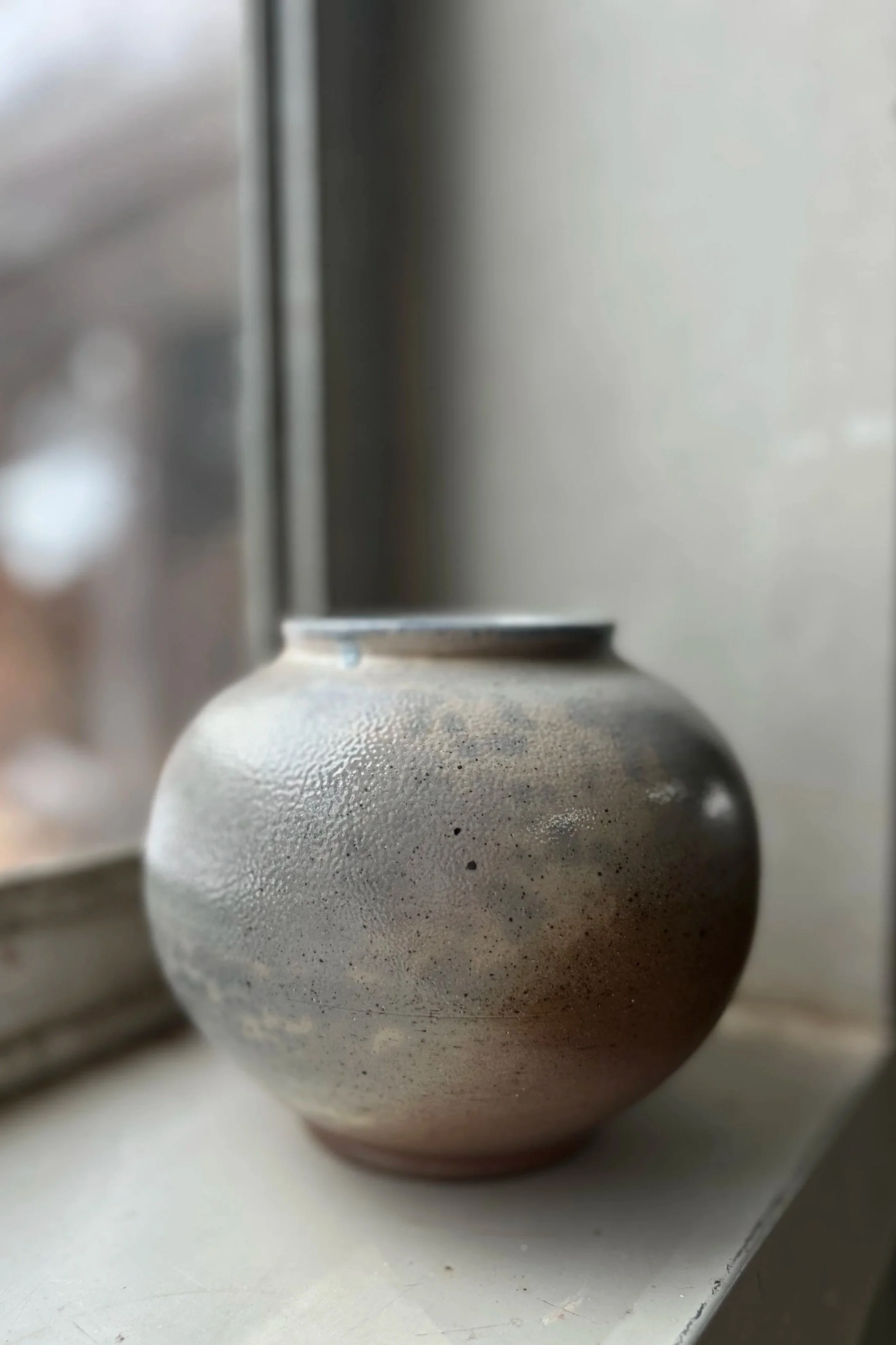 A textured soda-fired ceramic moon jar-inspired vase sitting on a white windowsill.