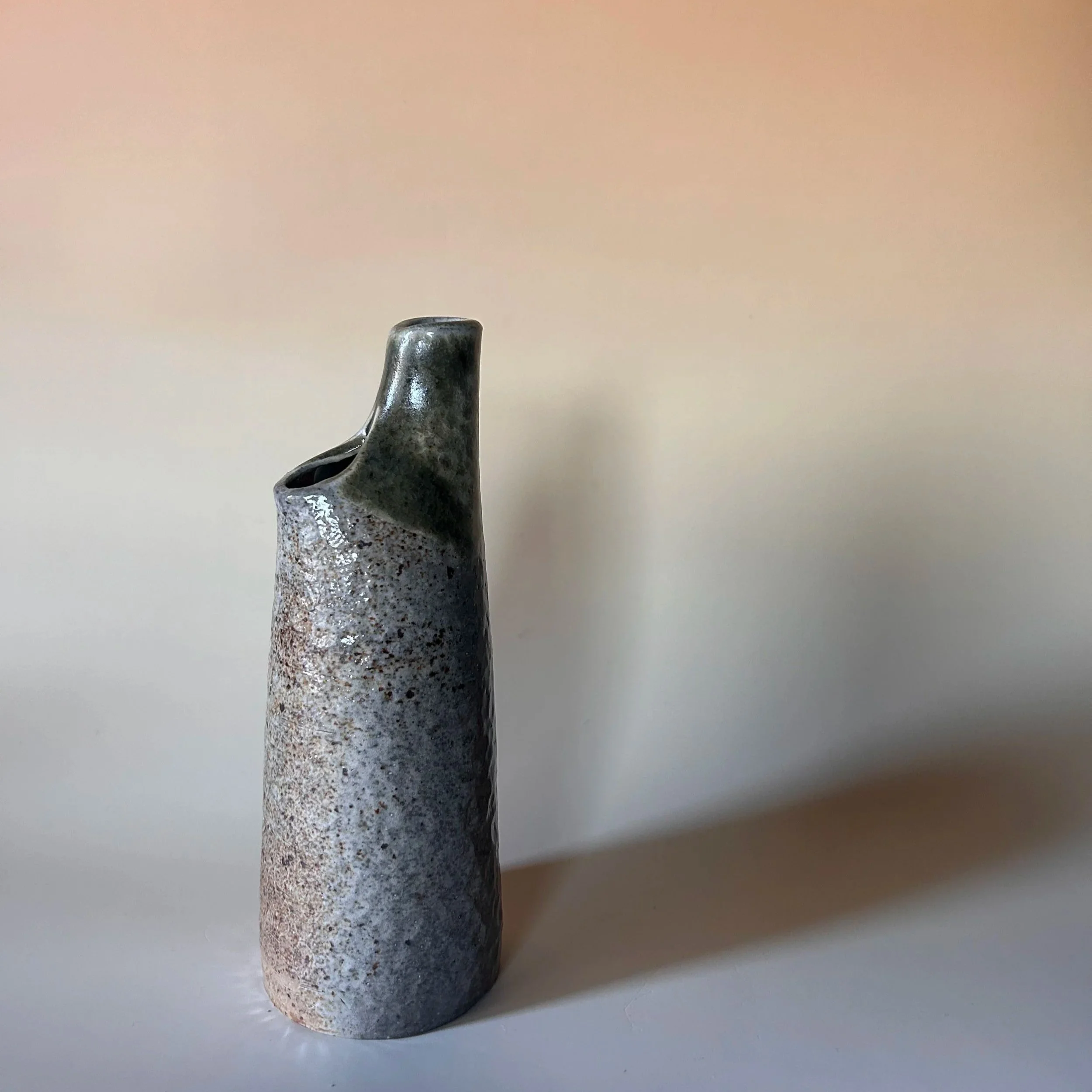 A tall ceramic soda fired bud vase with a textured, speckled gray surface and a glossy, darker neck on a plain white surface against a neutral background.