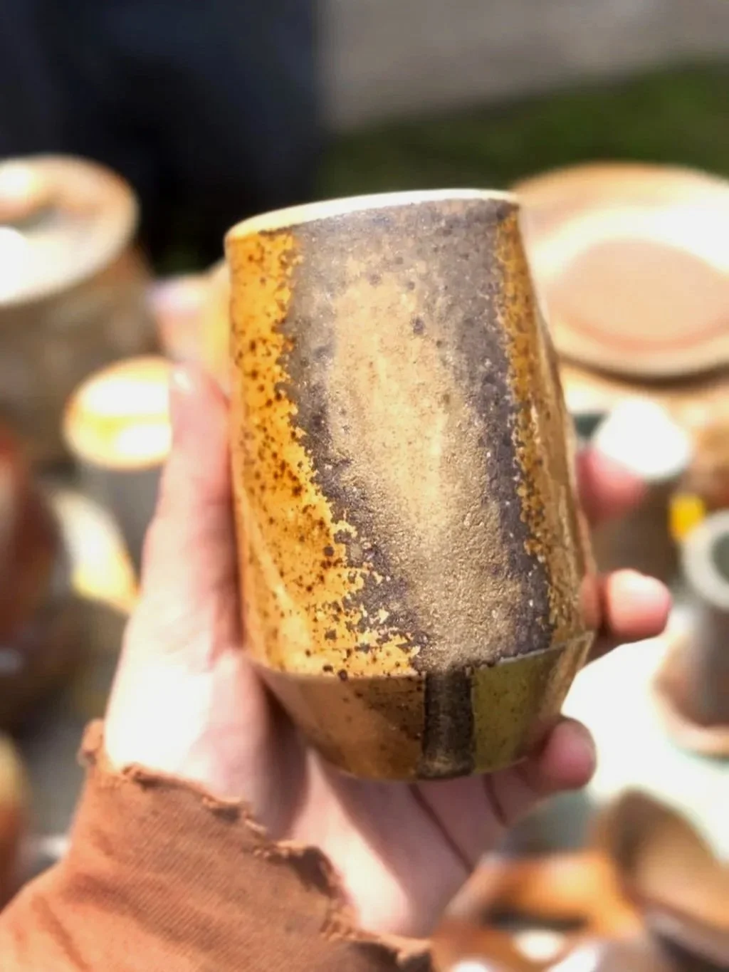Ceramic artist Amanda Cronin holding a wood fired angular cup with heavy ash glaze, in front of other similar mugs.