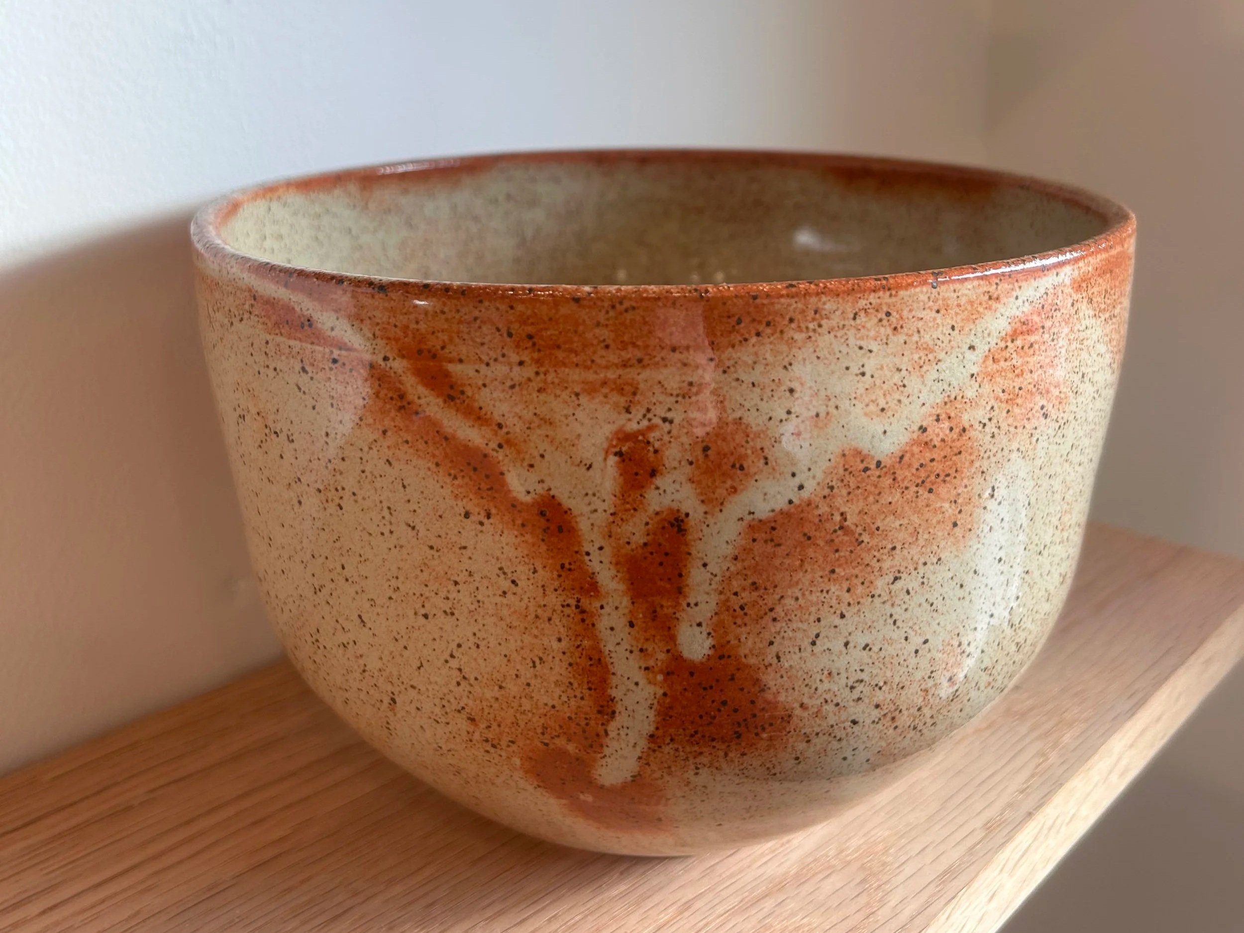 An electric-fired bowl with a shino-inspired cream-colored glaze with orange-brown glaze splashes sitting on a light wooden shelf.