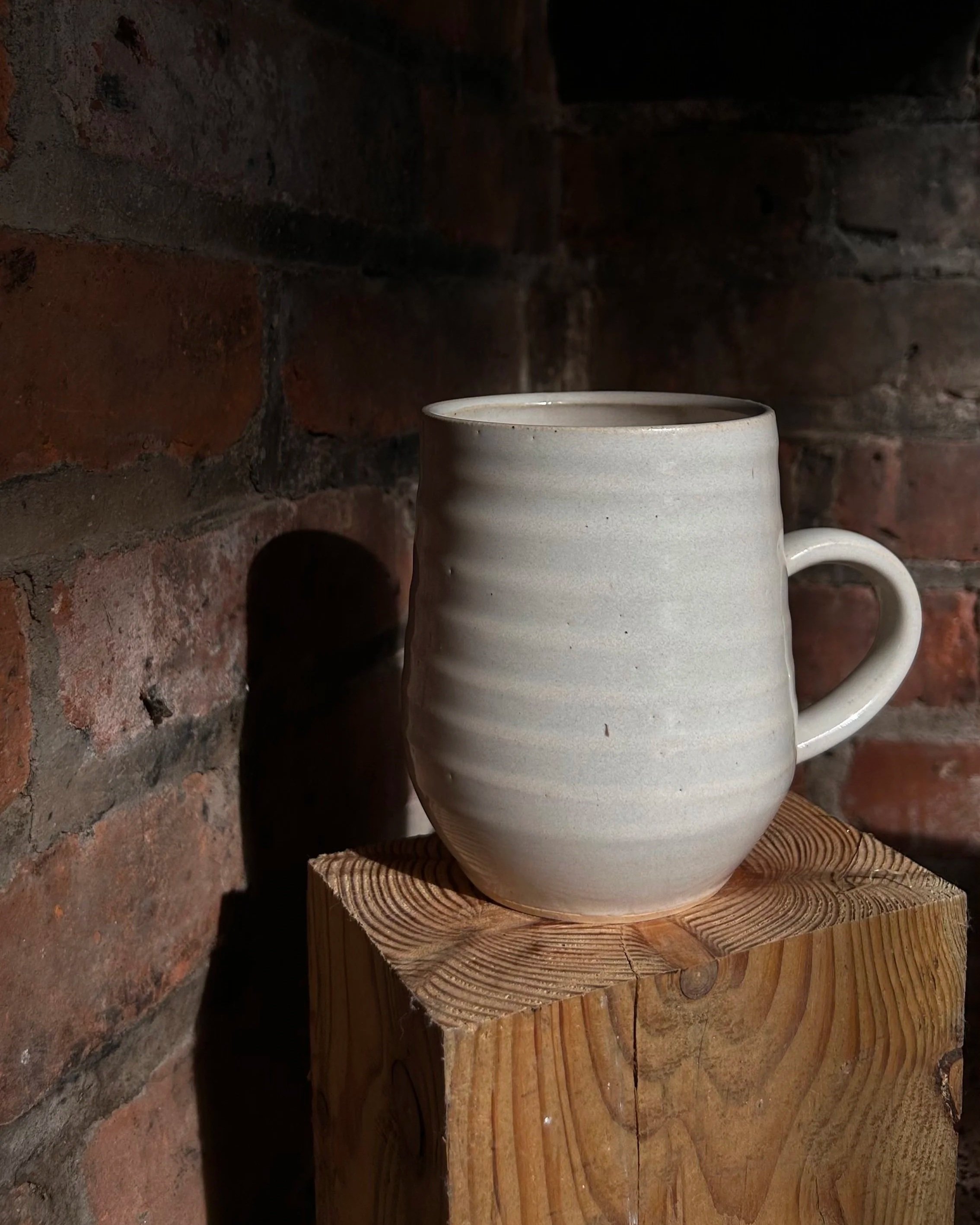 A white electric-fired ceramic mug with a textured, ribbed surface on a wooden block against a dark brick wall background.