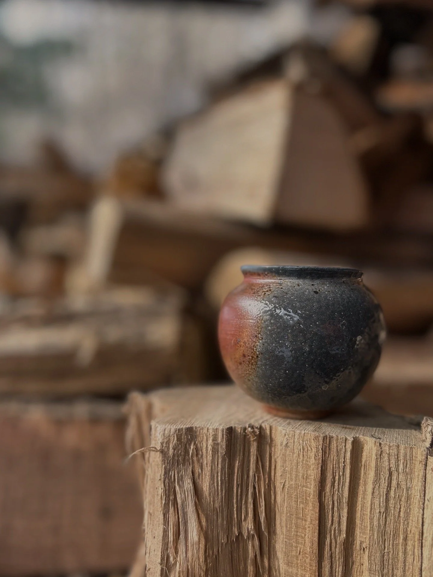 A mini soda-fired moon jar with blue, grey, and orange coloring, placed on a piece of wood with a blurred background of stacked wood logs.