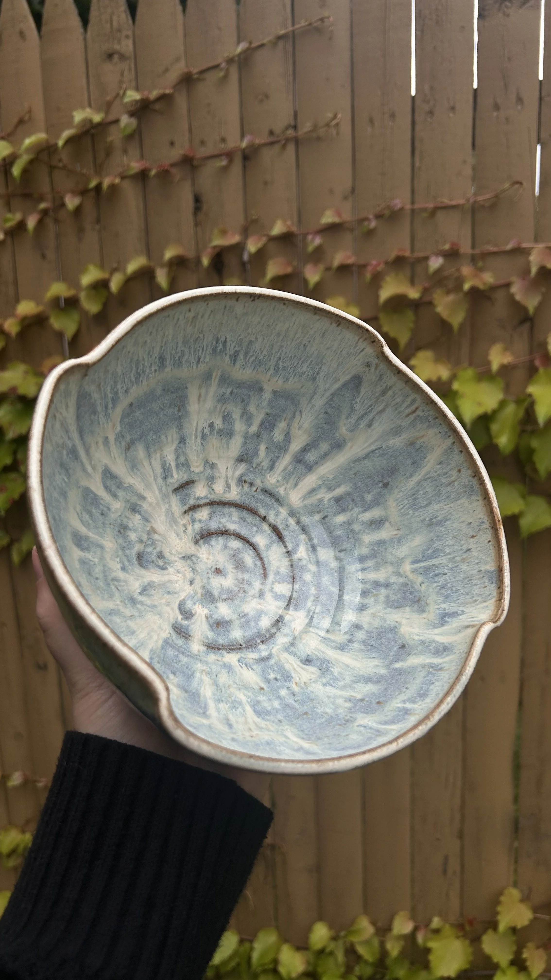 Ceramic artist Amanda Cronin holding an electric-fired ceramic bowl with an altered rim and sky blue and cream swirling pattern inside, outdoors with a wooden fence and climbing ivy in the background.