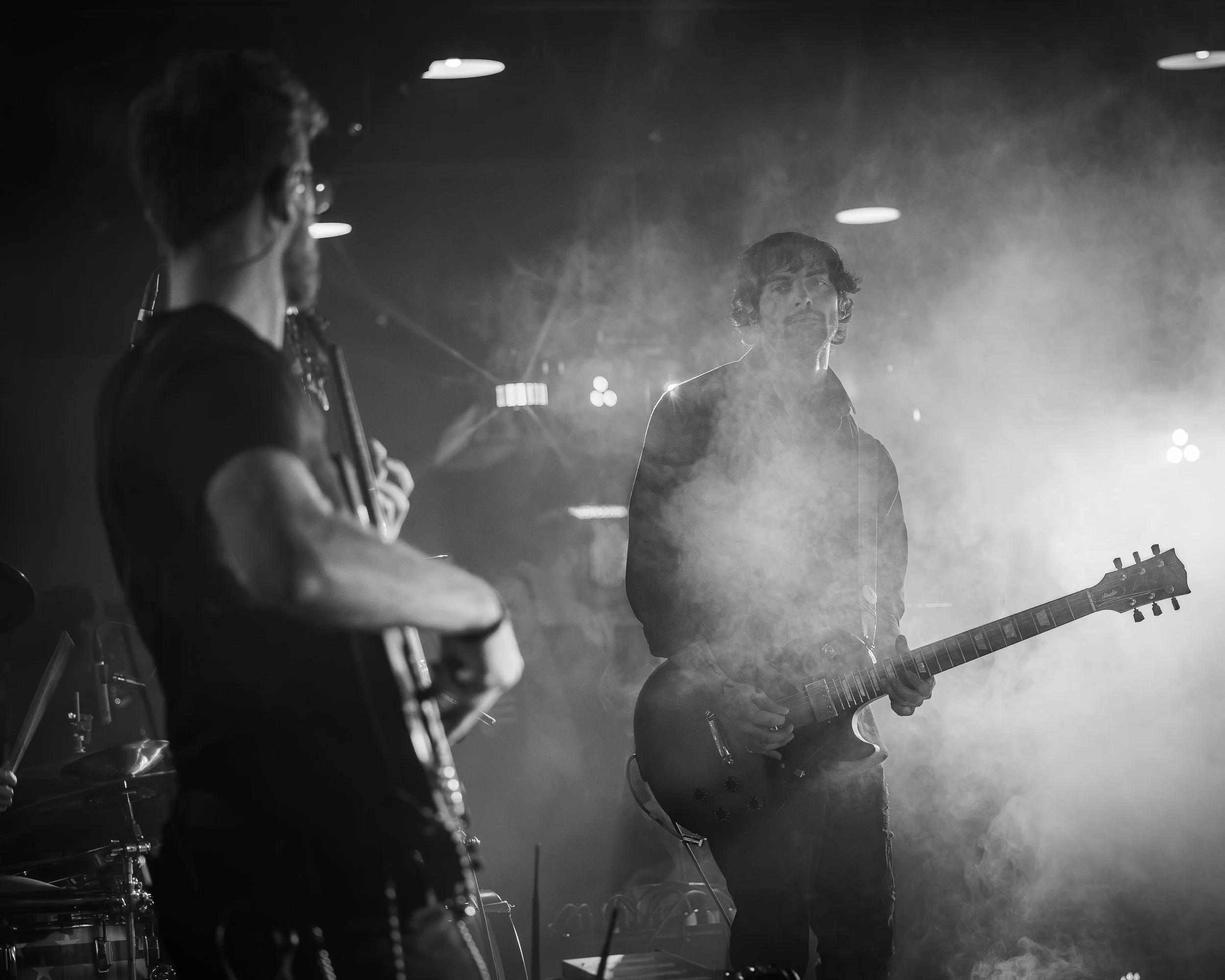Two musicians performing on stage with a smoky background, one playing an electric bass guitar and the other playing an electric guitar.