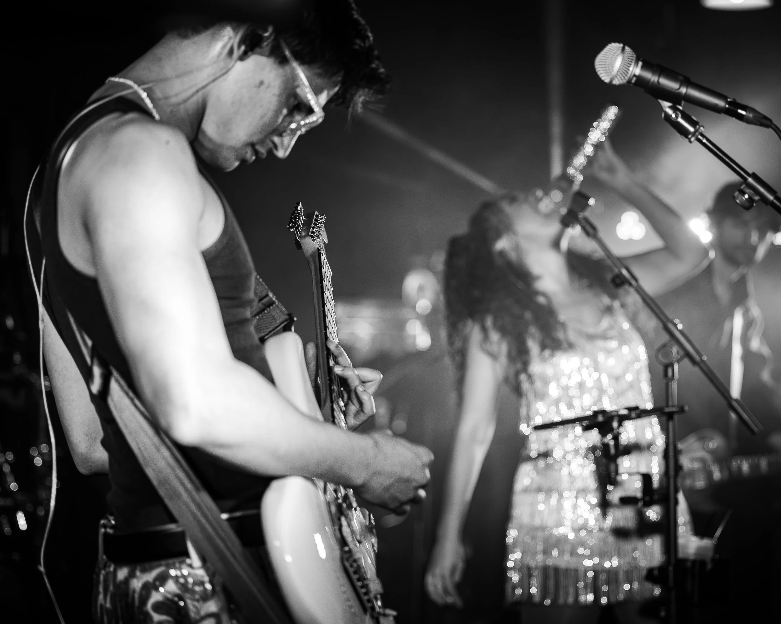 Musicians performing on stage, with one person playing electric guitar and woman singing into a microphone, in black and white.