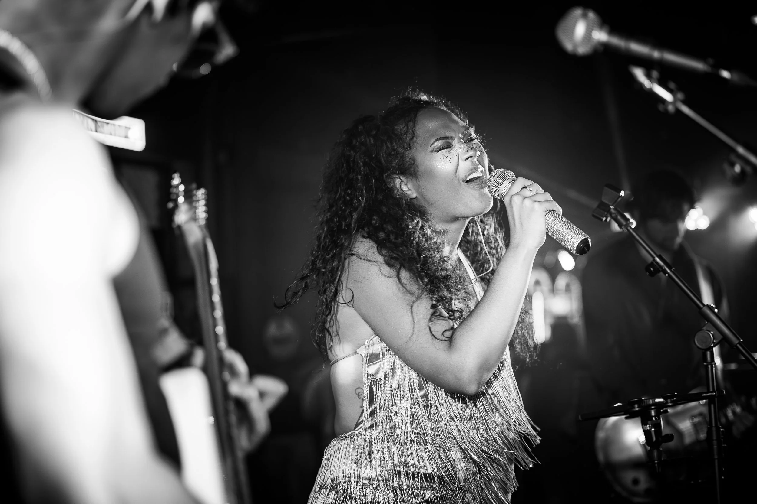 A woman with curly hair singing passionately into a microphone on stage, with a male musician in the background and musical instruments around.