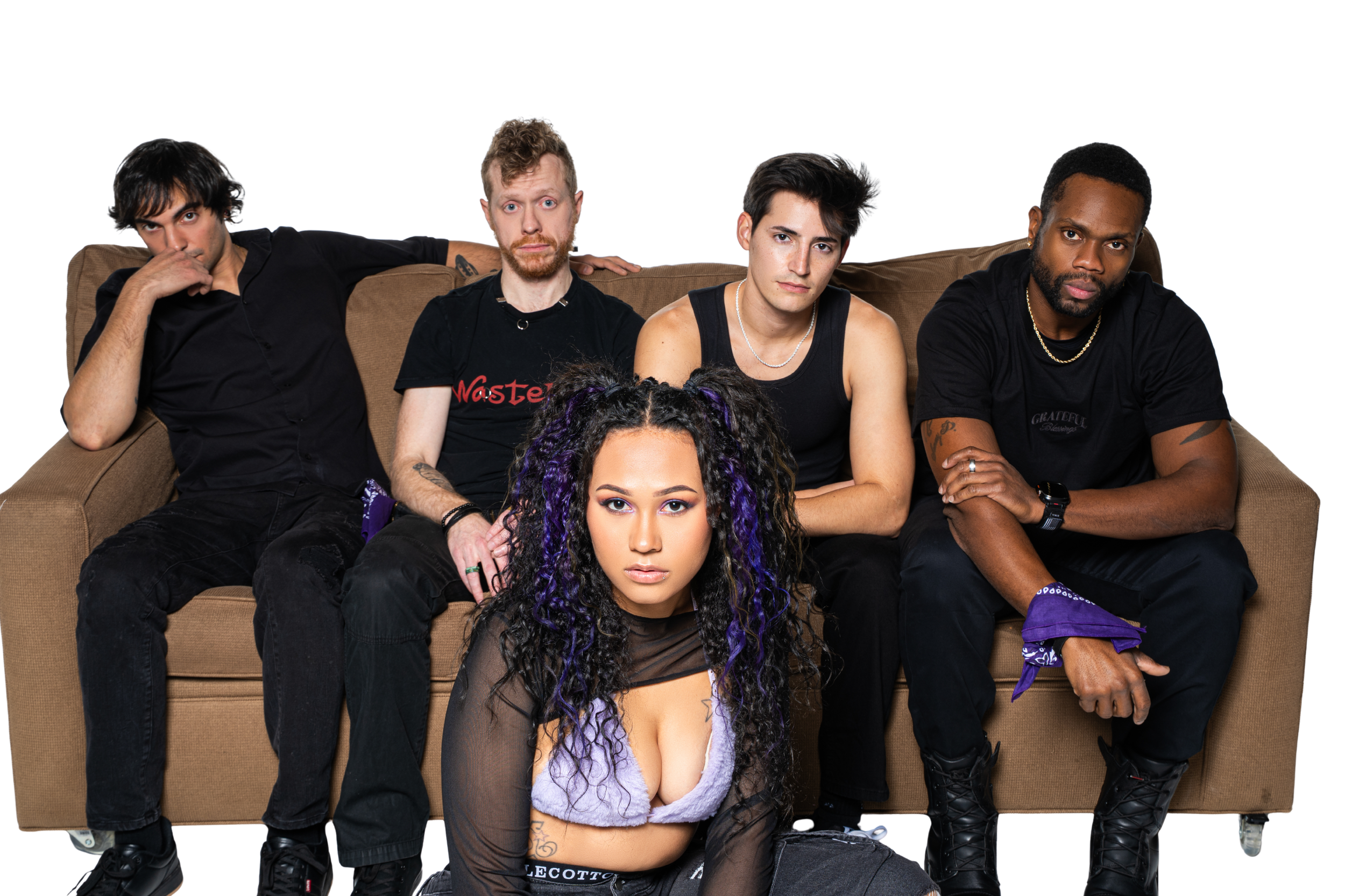 Group of five diverse young adults sitting on a beige sofa, with one woman kneeling on the floor in front. The woman has curly purple and black hair and wears a sheer black top over a lavender bra, with various tattoos visible on her arms. The men wear dark casual clothing, with one wearing a tank top, and all look directly at the camera with serious expressions.