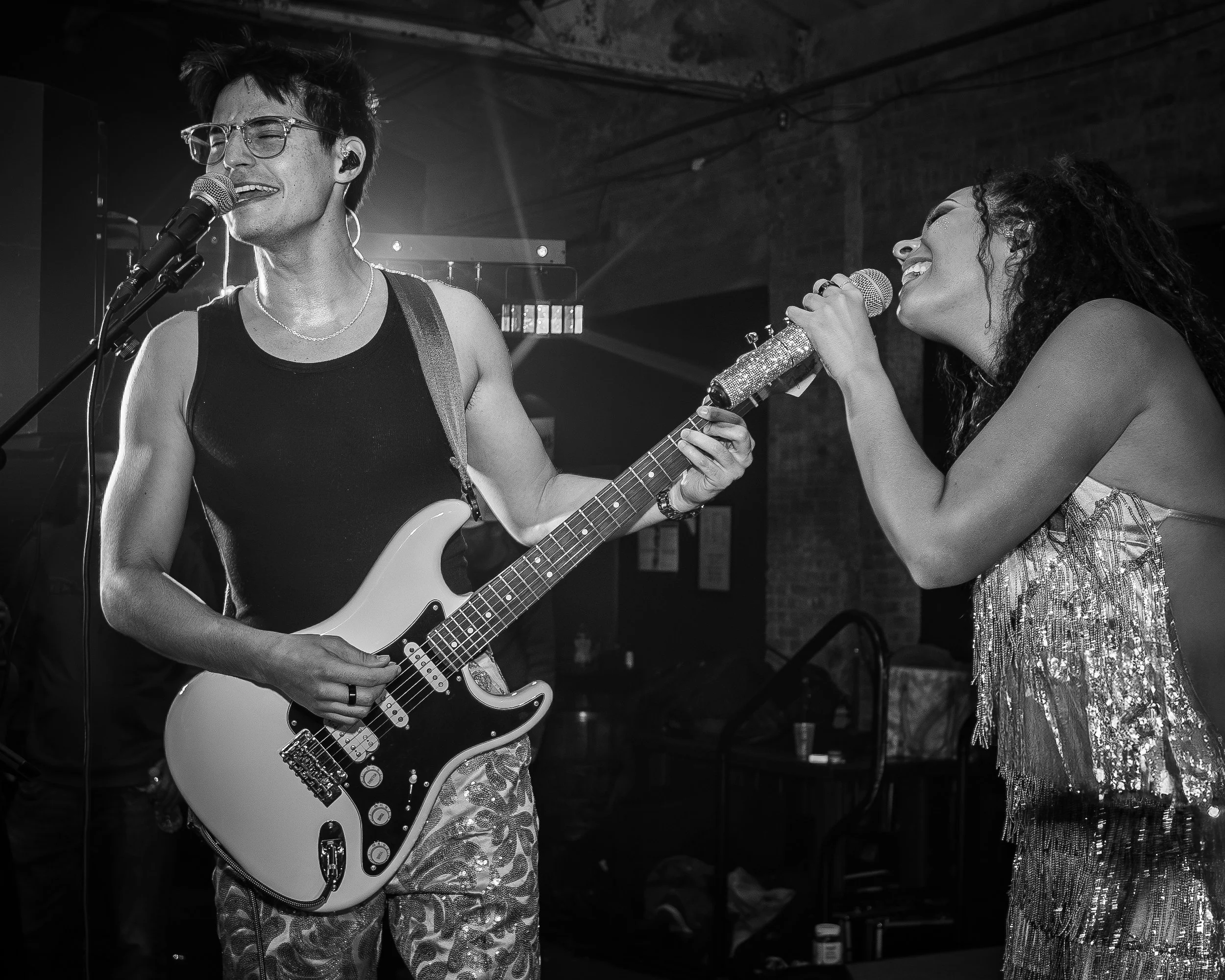 A man playing electric guitar and singing into a microphone while a woman sings into her microphone during a live performance in a dimly lit venue.