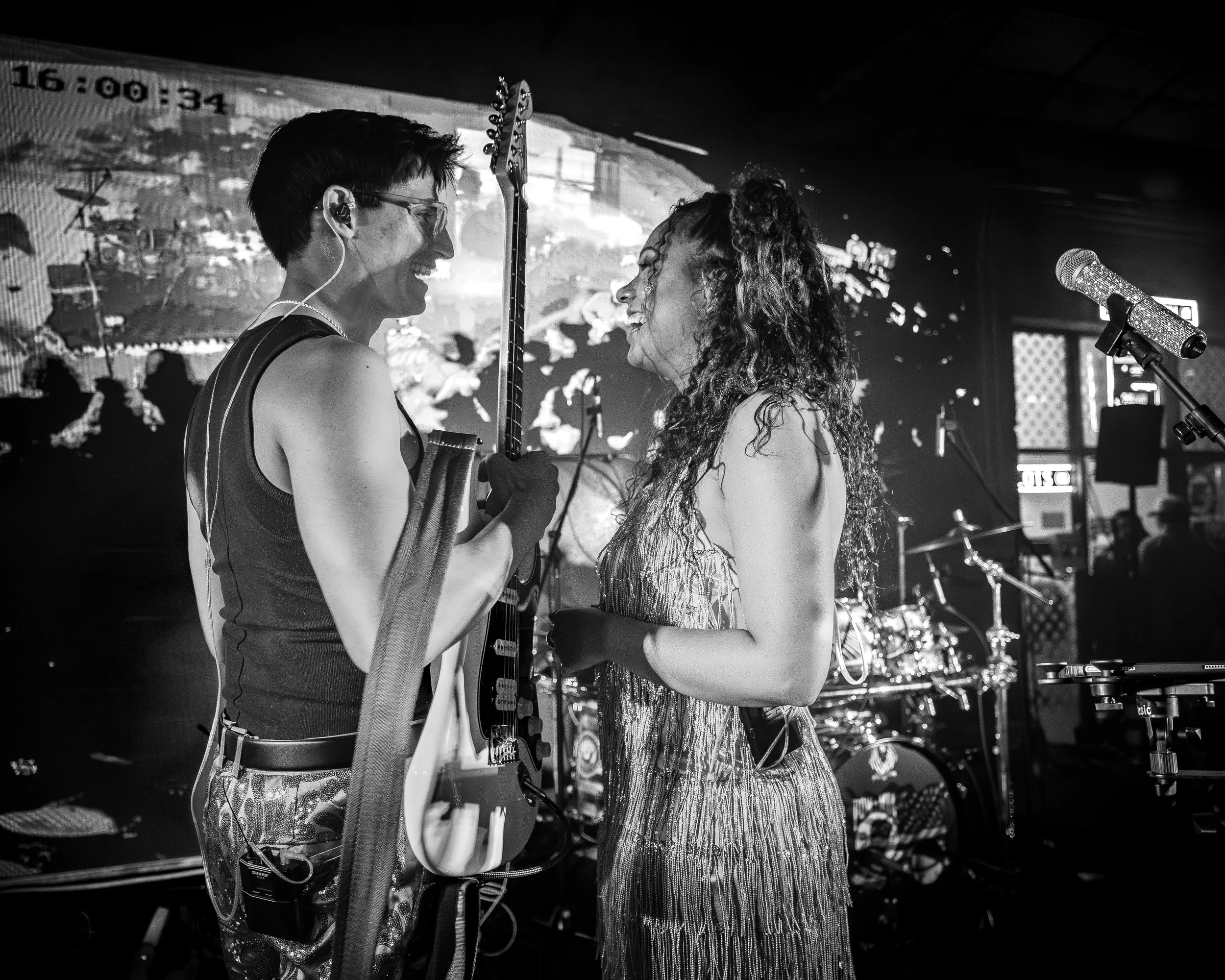 Two musicians, a man and a woman, share a joyful moment on stage during a live performance, with musical instruments and stage equipment in the background.