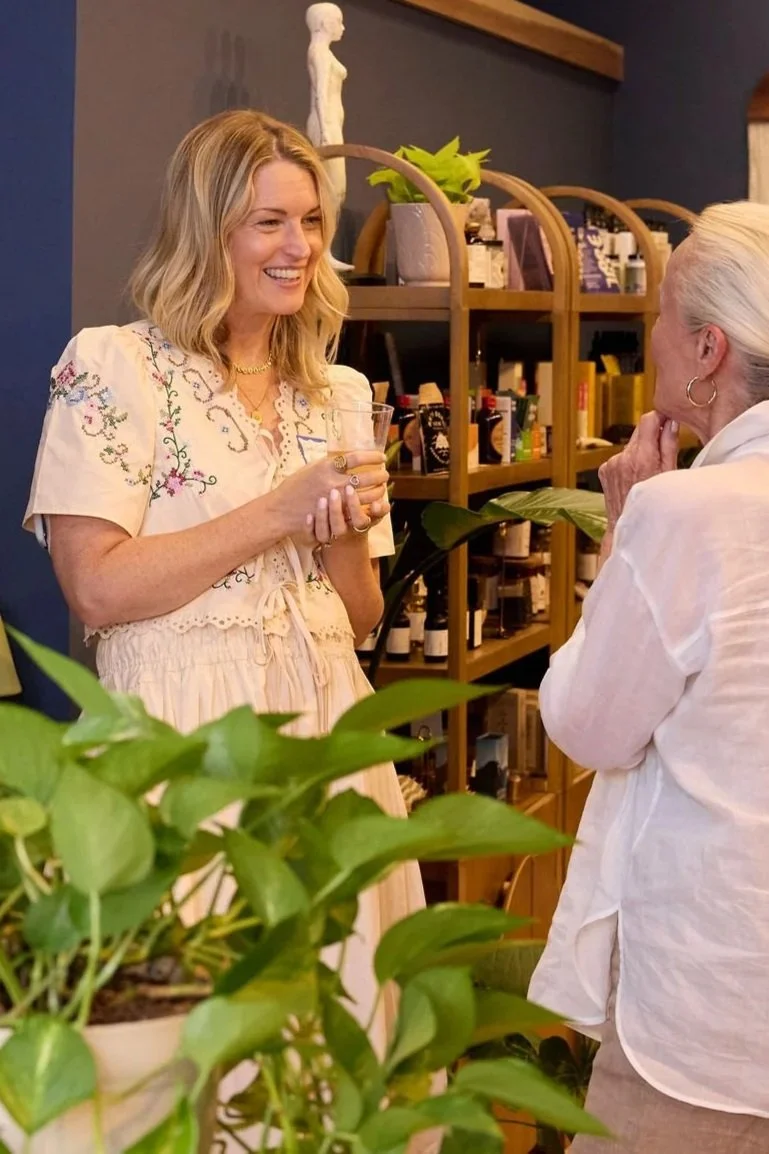 Two women are having a conversation in a cozy room filled with plants and bookshelves. One woman has blonde wavy hair and is smiling, holding a glass, while the other woman has white hair and is wearing earrings, listening attentively.