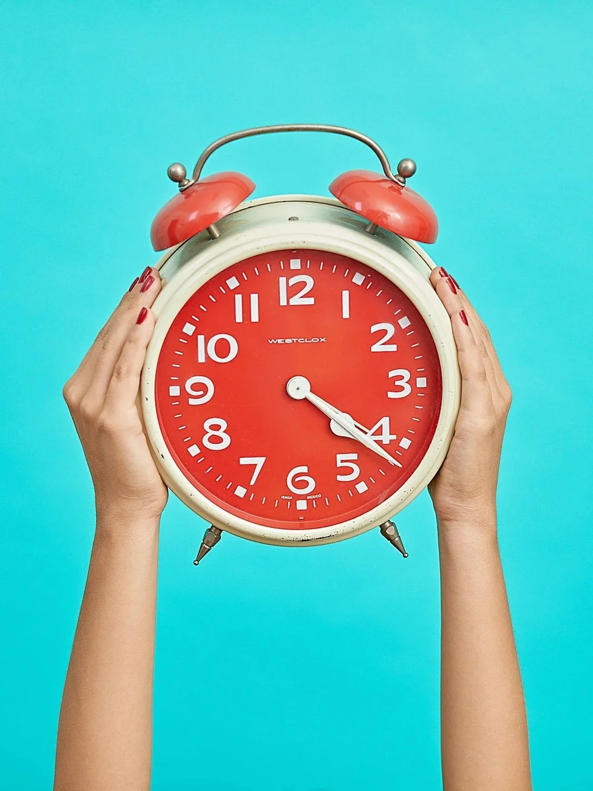 Two hands holding a vintage red alarm clock with a bright turquoise background.