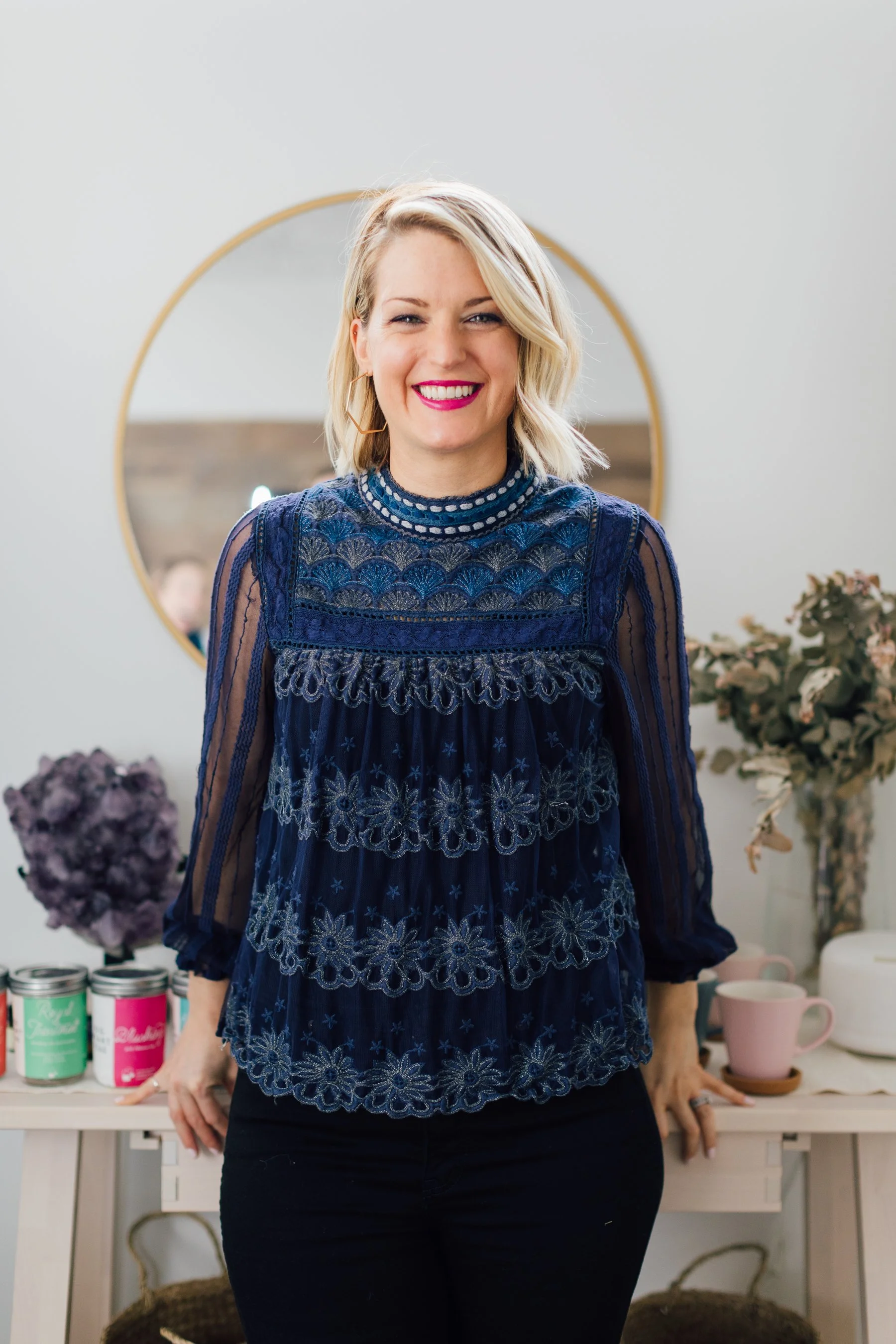 A woman with blonde, wavy hair and pink lipstick smiling at the camera in a room decorated with a round mirror, plants, and pink and white mugs.