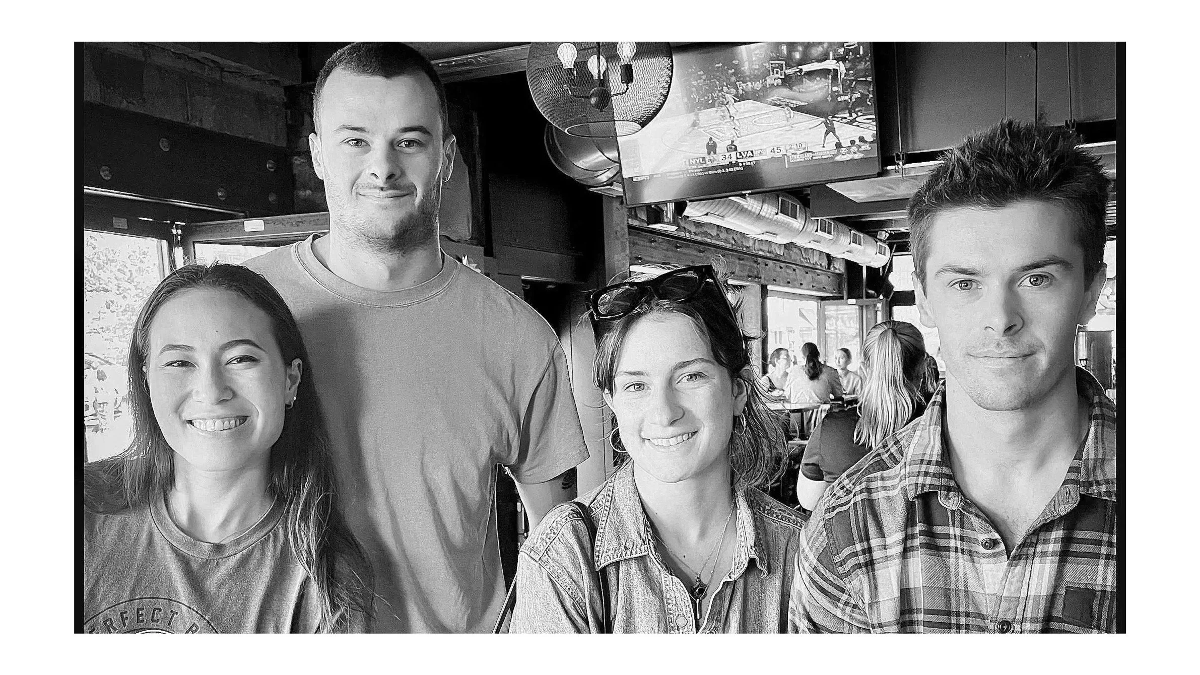 Four young adults, two men and two women, smiling at the camera inside a bar or restaurant with a TV screen showing a basketball game in the background.