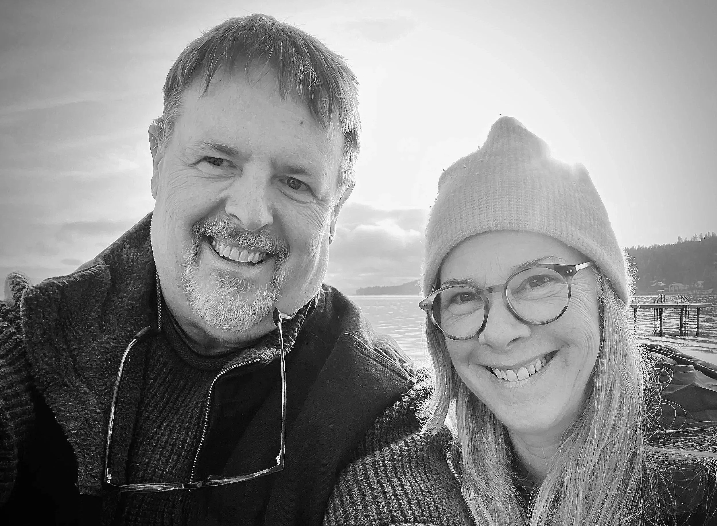 A smiling man and woman taking a selfie outdoors near a body of water, with trees and a dock in the background, during what appears to be a sunny day.