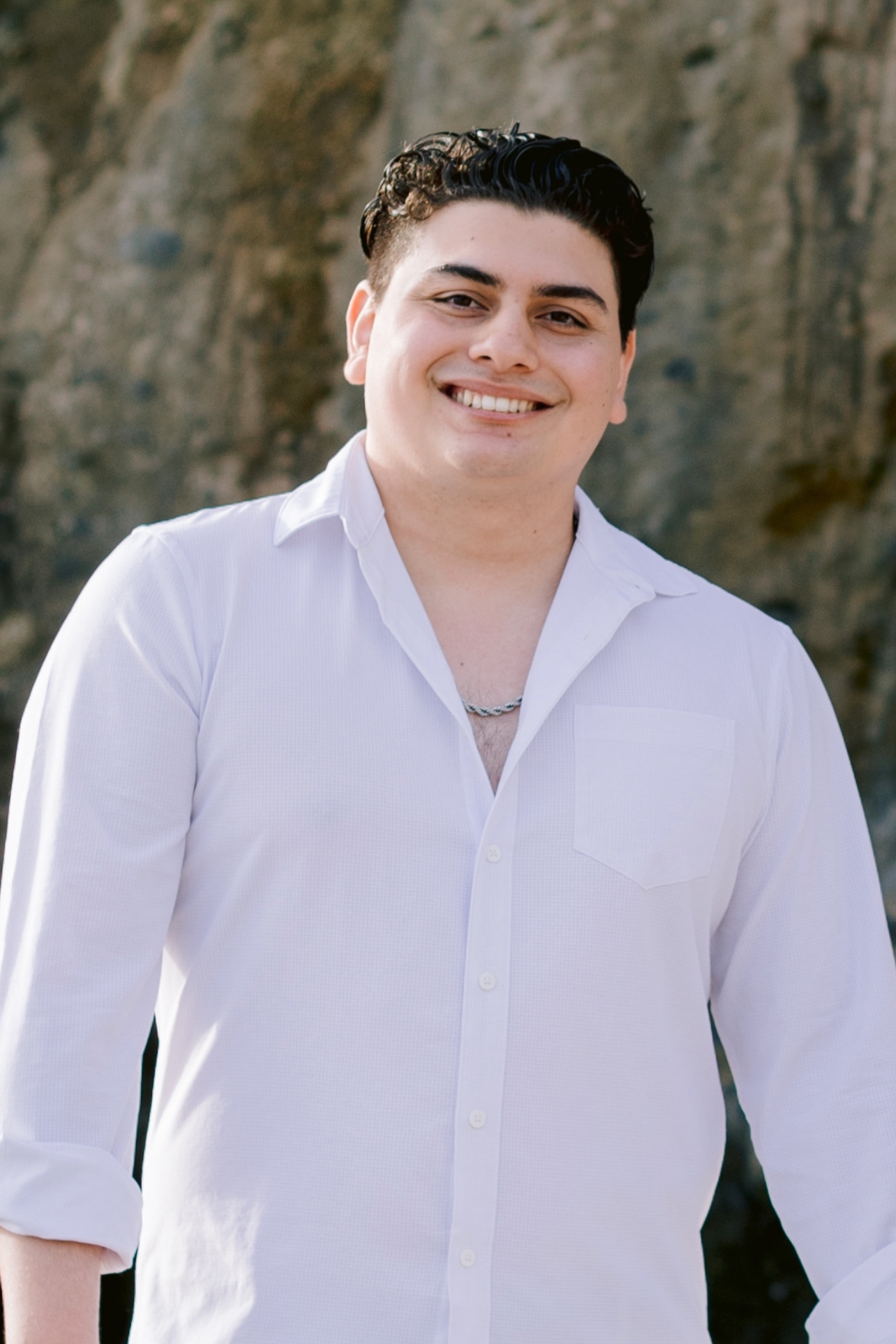 A young man smiling outdoors with a rocky background, wearing a white button-up shirt and a necklace.