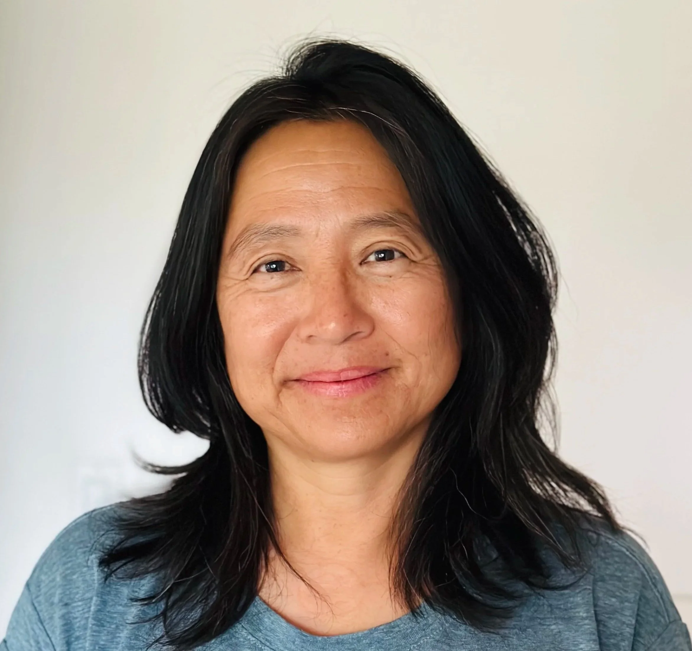 A woman with shoulder-length dark hair, smiling, wearing a gray shirt against a plain light-colored background.