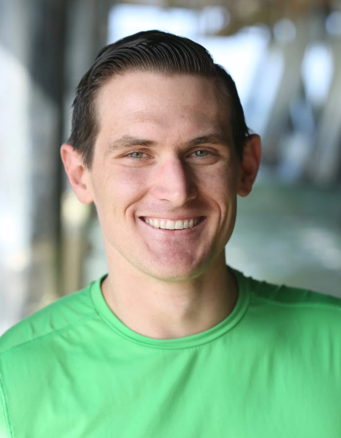 Close-up portrait of a young man smiling, with short dark hair, light skin, wearing a green shirt, outdoors with blurred trees in the background.