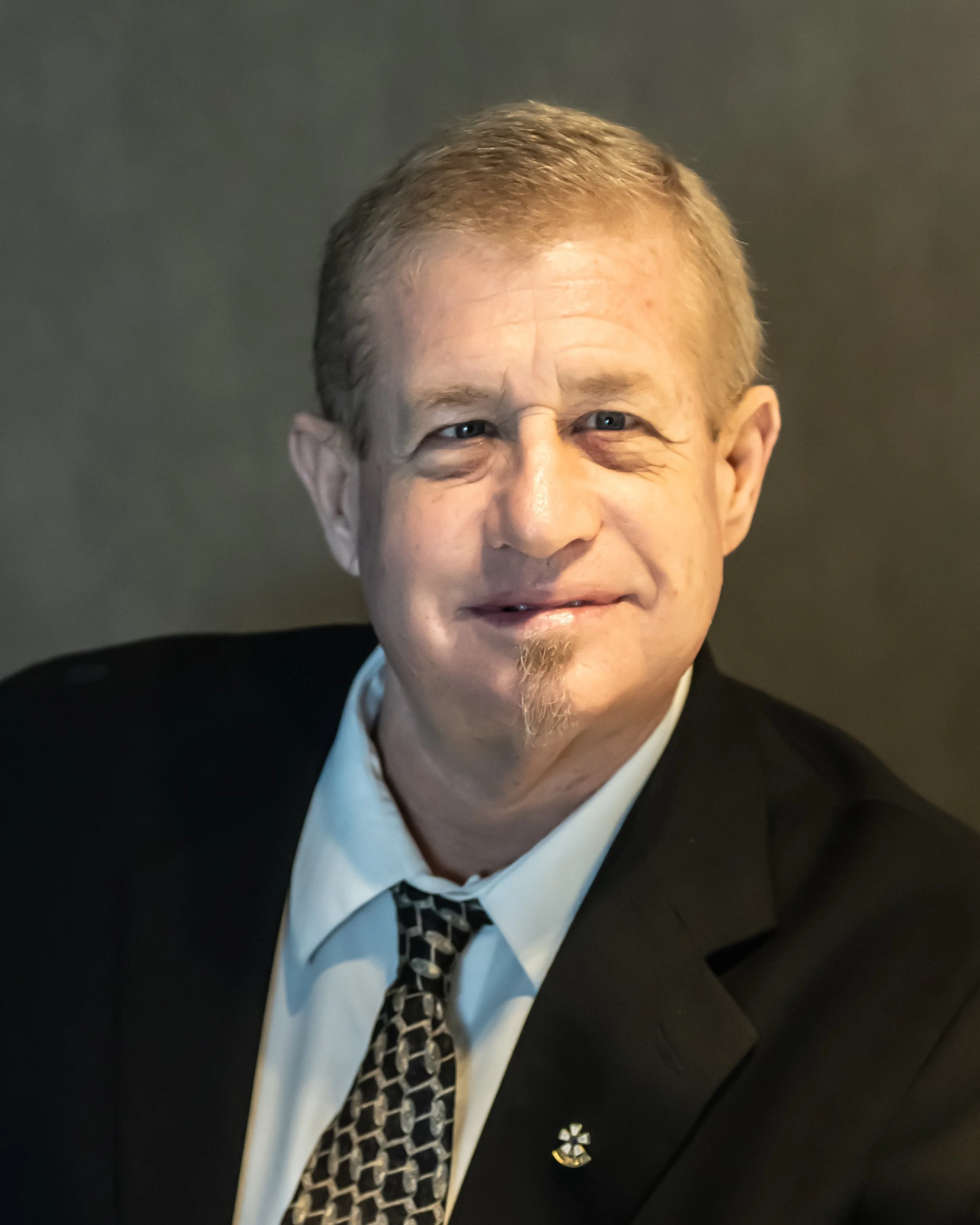 A middle-aged man with blond hair and a goatee, wearing a black suit, white shirt, and patterned tie, smiling at the camera against a dark background.