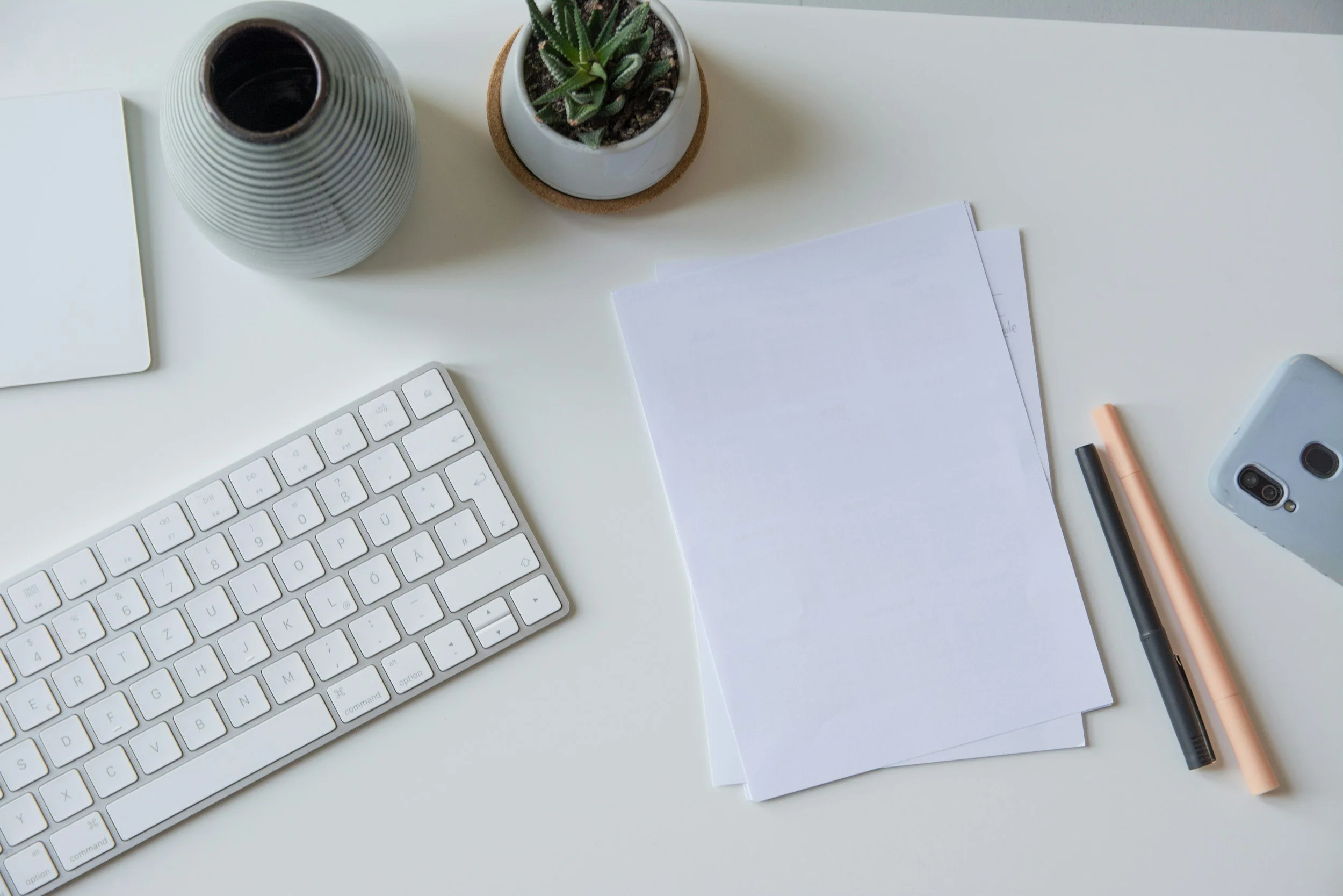 A white desk with a wireless keyboard, stacked papers, a grey phone, a black pen, a peach-colored pen, a small potted cactus, and a grey-striped vase.