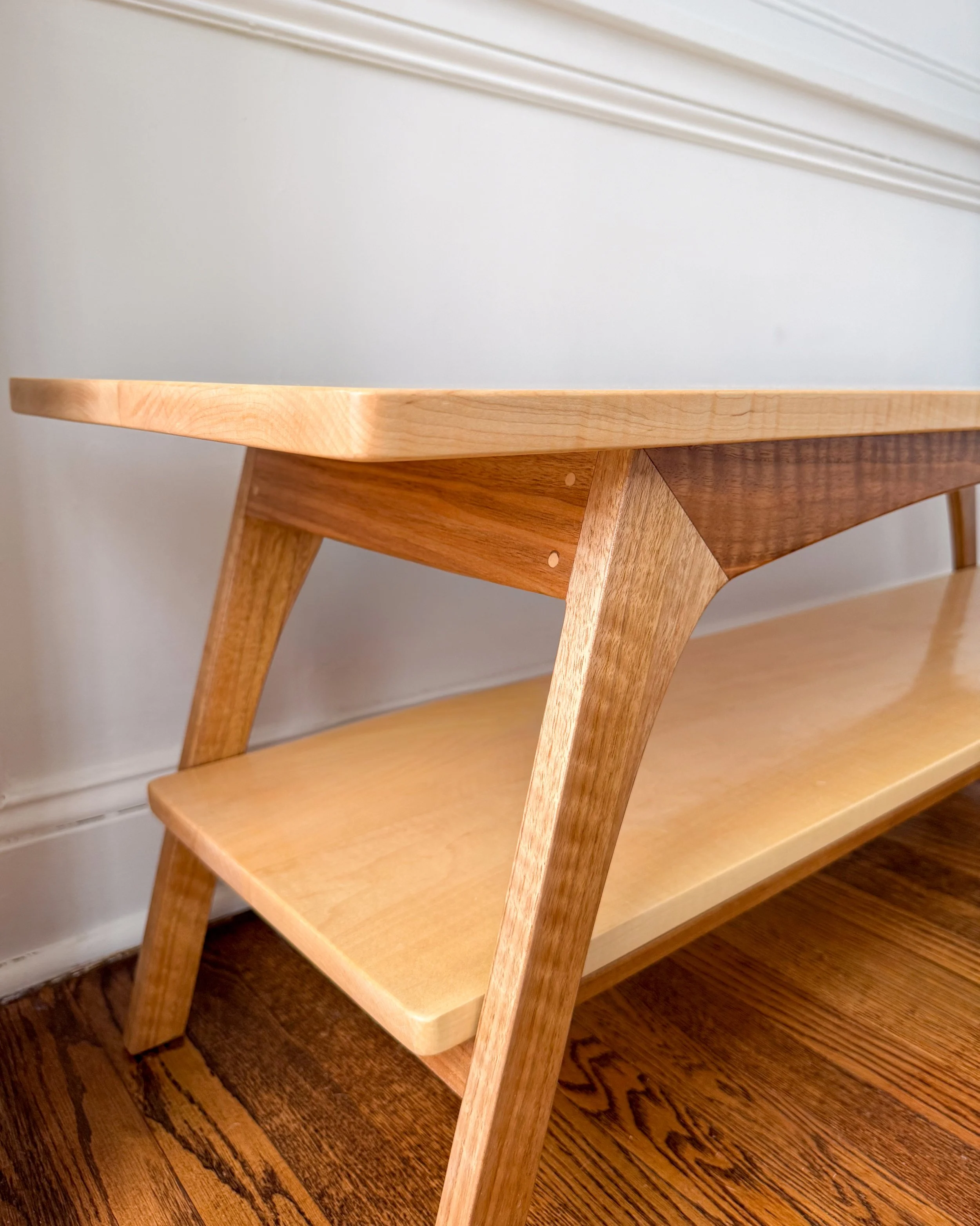 Close-up of a modern wooden bench with a light wood top and a lower shelf, against a white wall with molding.