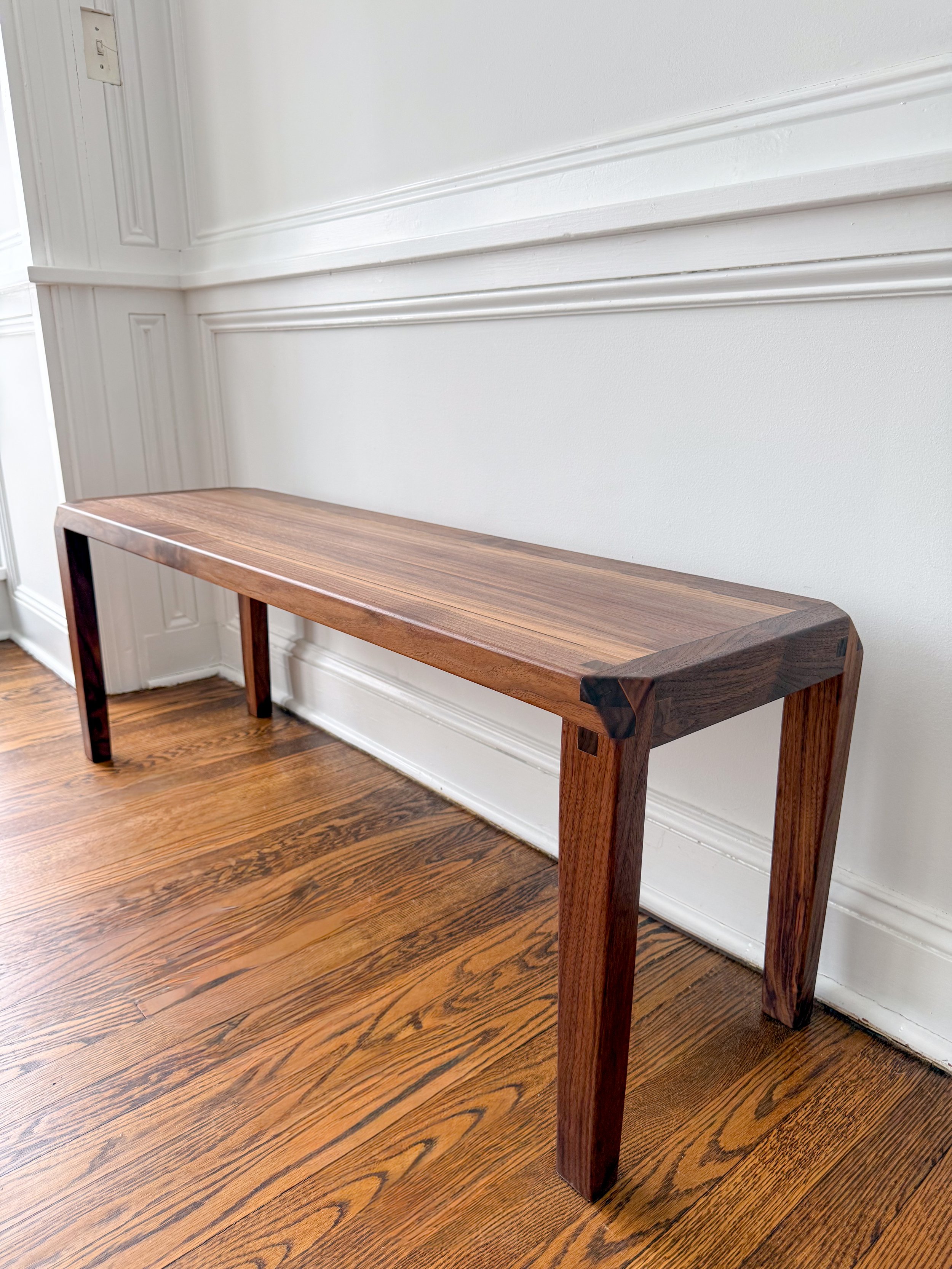 A wooden bench with a polished wood top and matching wooden legs in a room with hardwood flooring and white wainscoting.