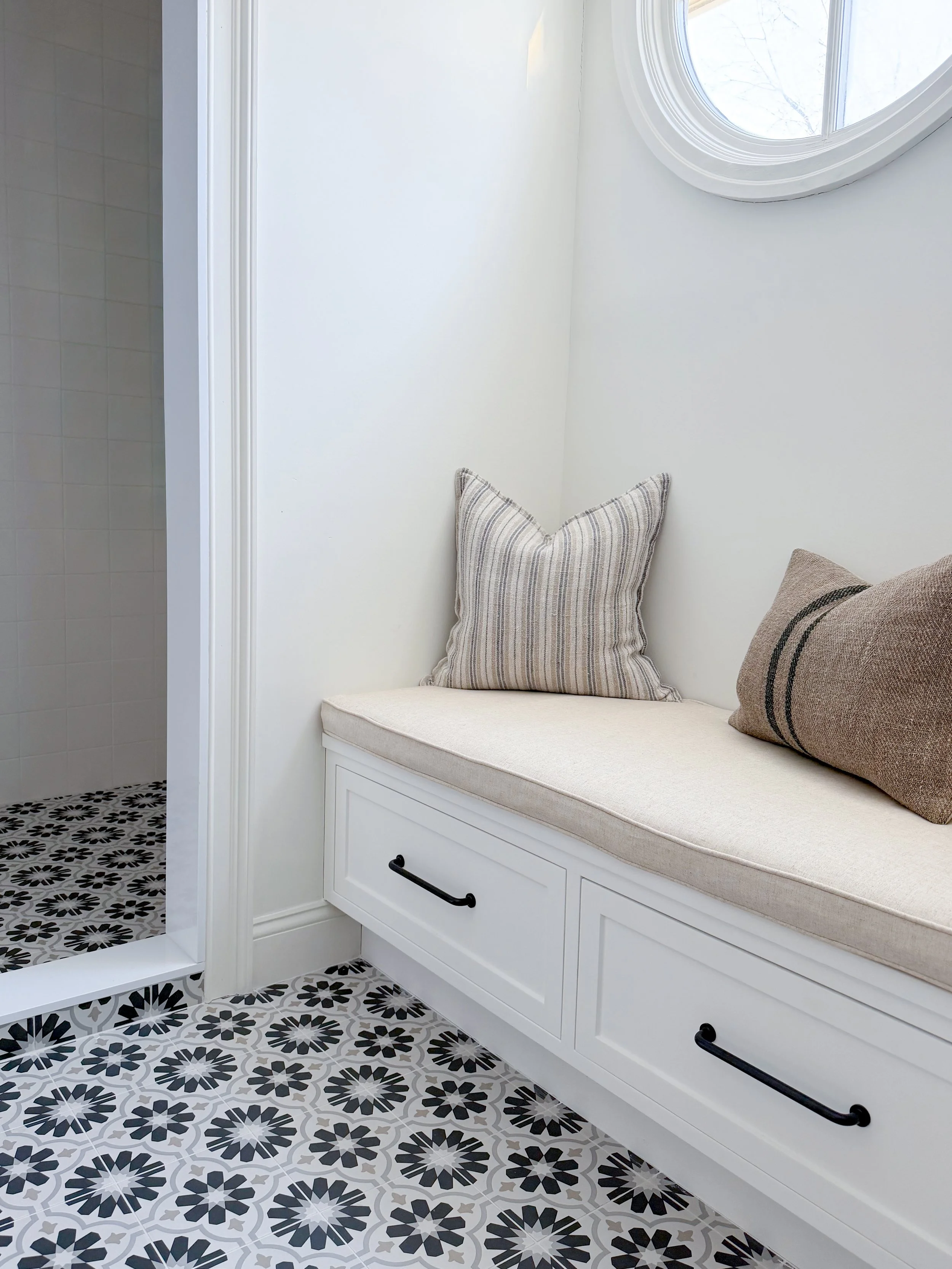 A cozy window seat with pillows in a bathroom, next to a walk-in shower with black and white patterned tile flooring.