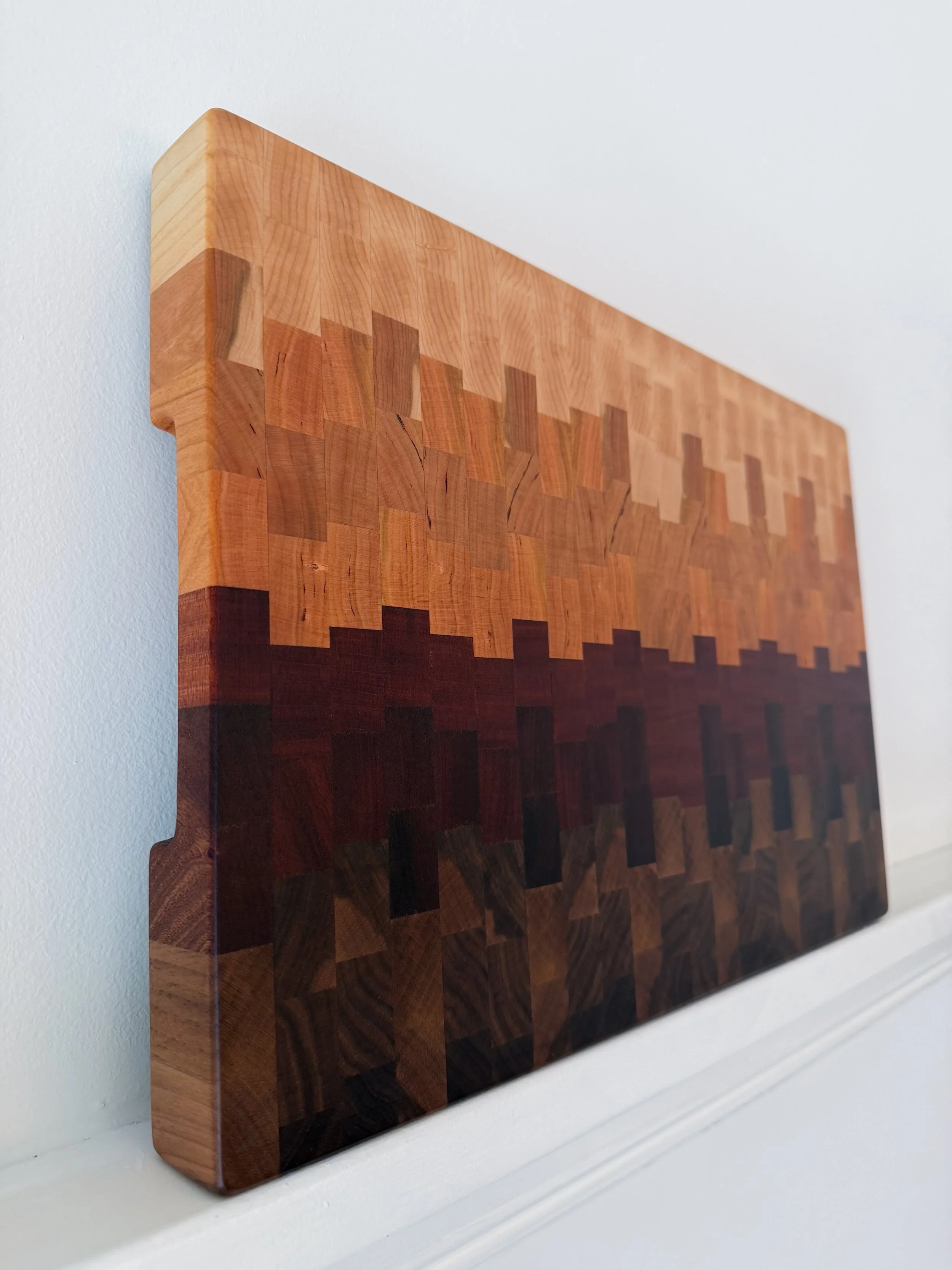 A wooden art piece with a pixelated landscape design, featuring a gradation of colors from light to dark, mounted on a white wall.