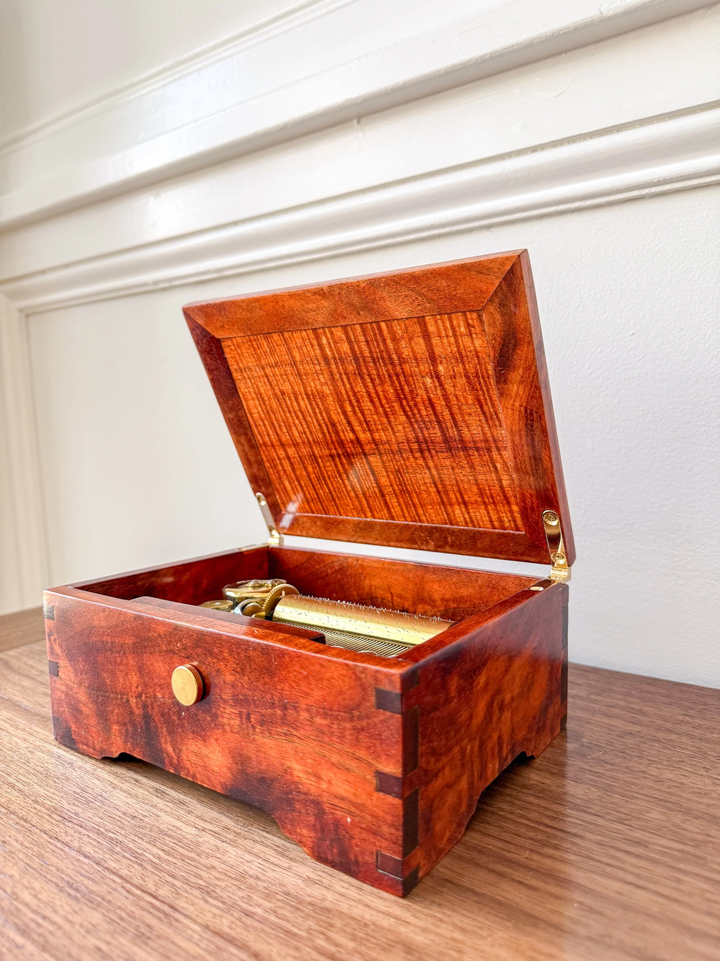 Open wooden box with a brass music box mechanism inside, placed on a wooden surface against a white wall with decorative molding.