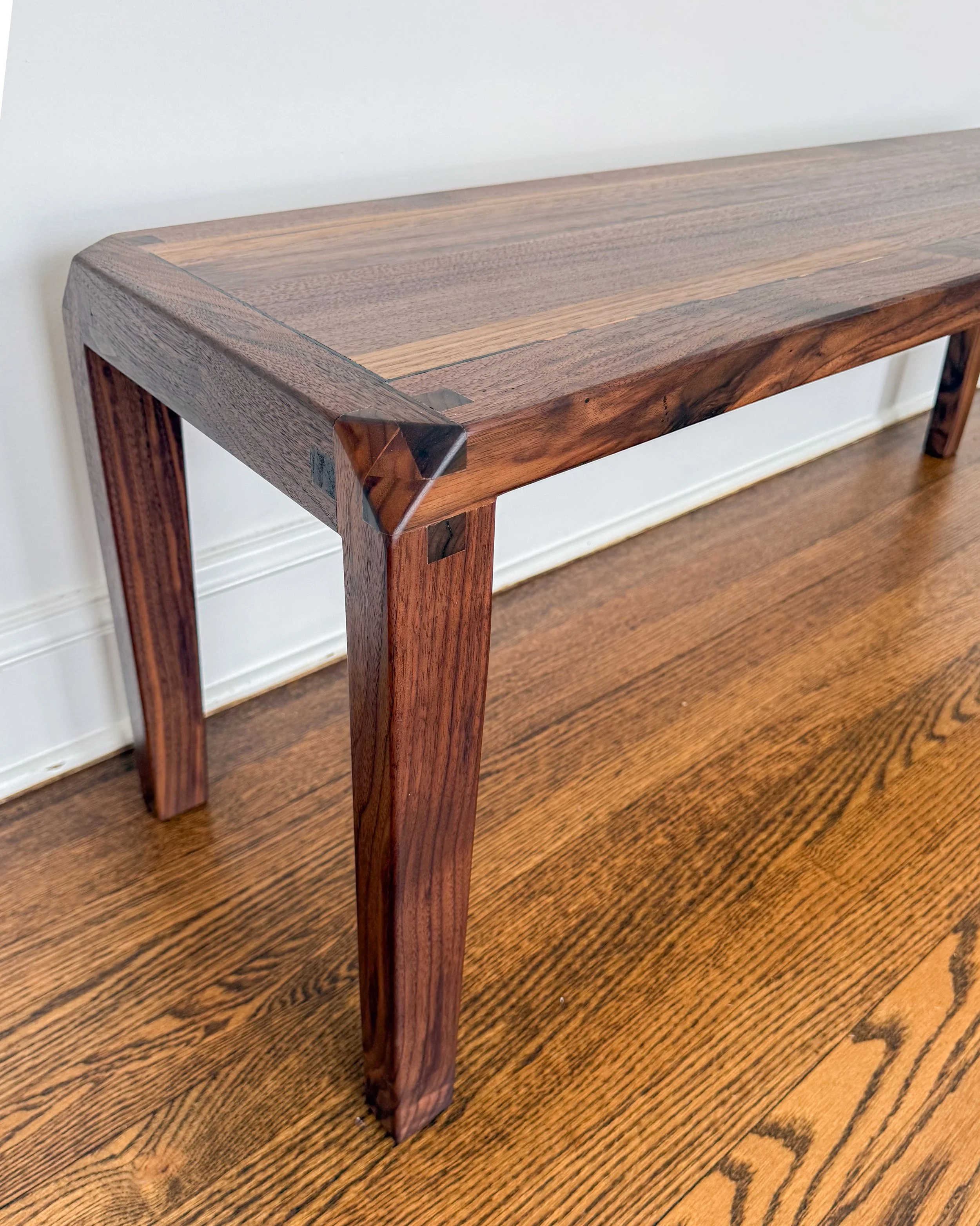 Close-up of a wooden table with a smooth surface, showcasing the natural wood grain and a polished finish, placed on a hardwood floor against a white wall.