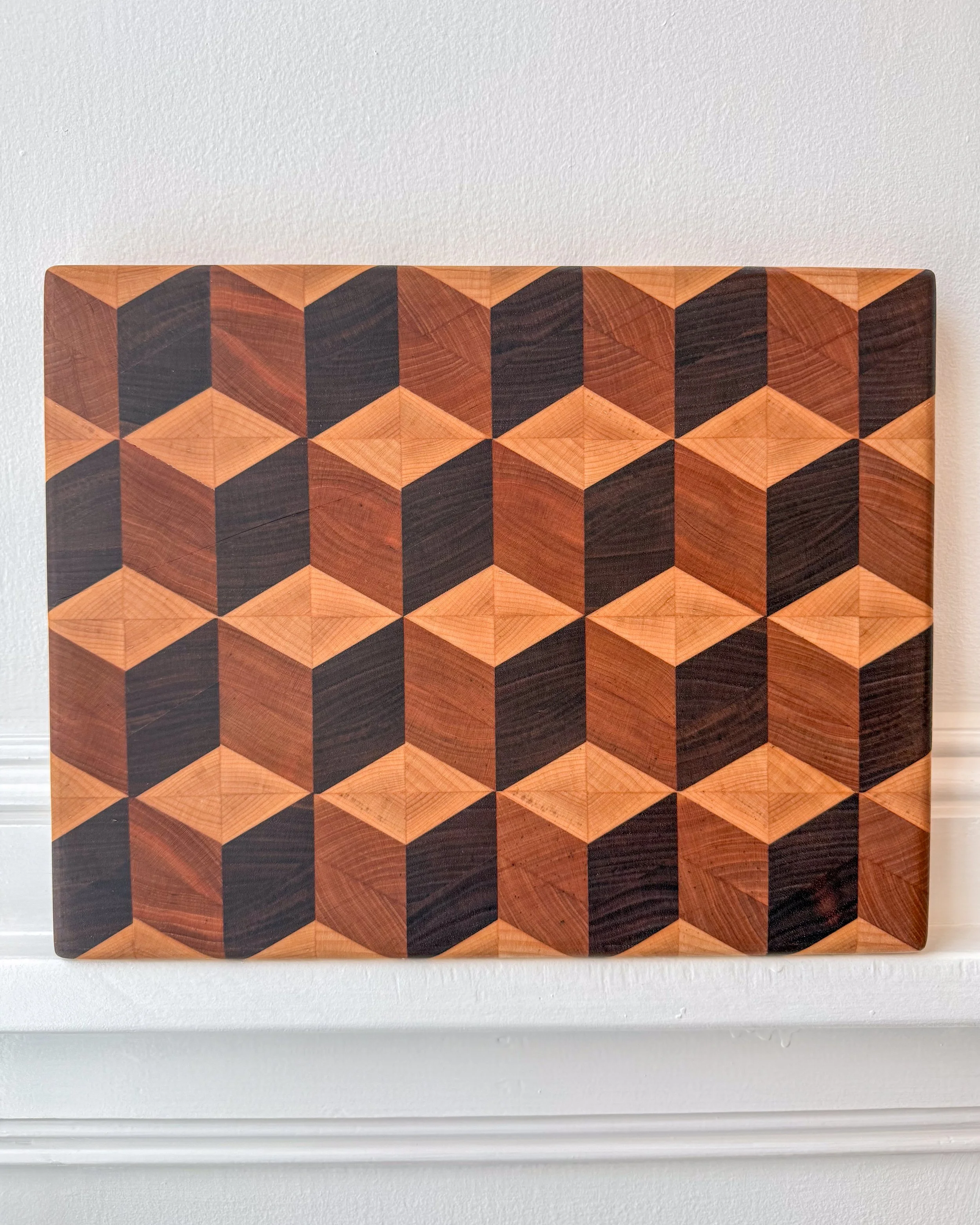 A wooden geometric patterned art piece with cube illusion, made of multiple wood tones, mounted on a white wall.