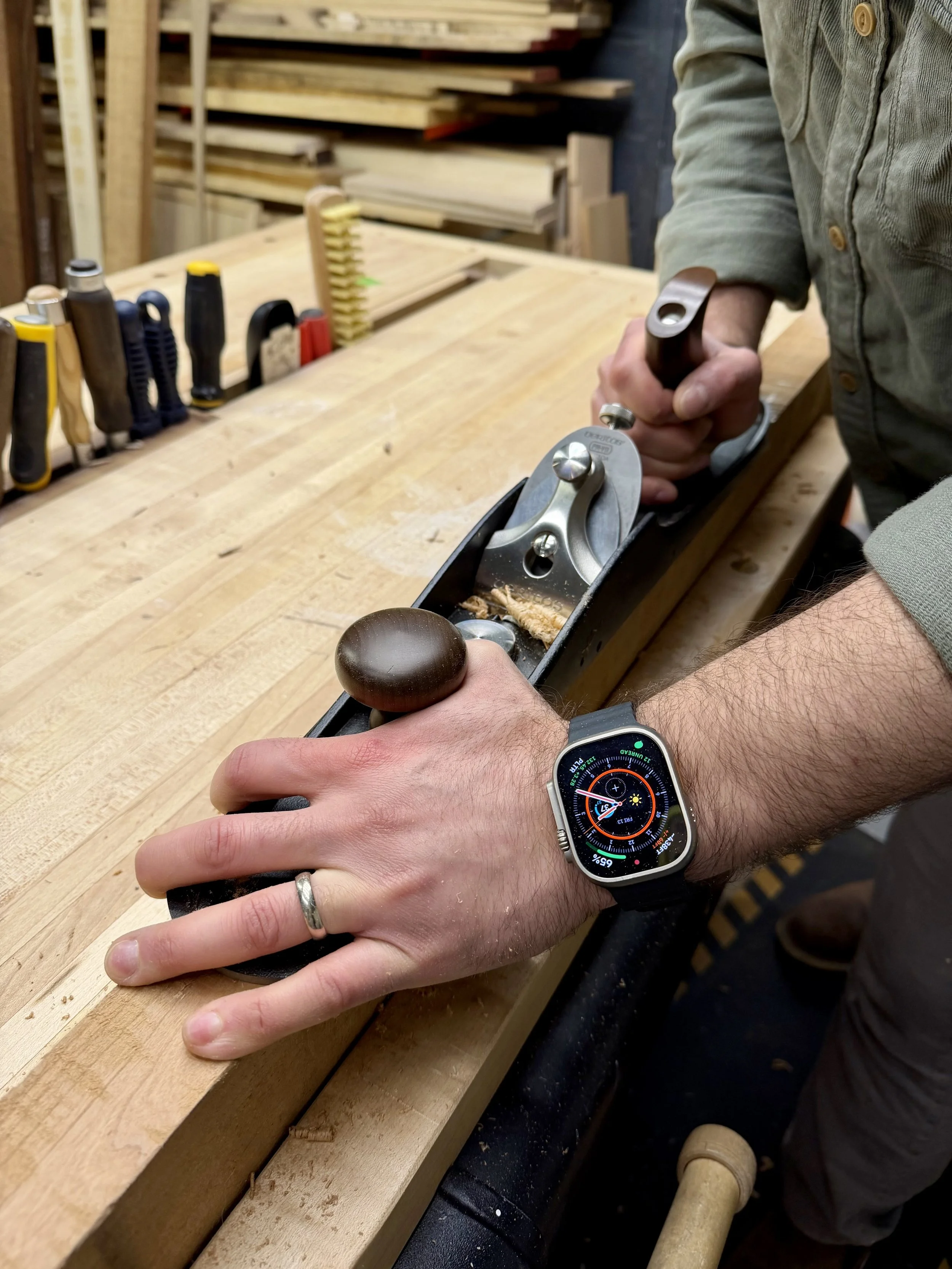 Person using a hand plane tool to smooth a piece of wood on a workbench in a woodworking shop.
