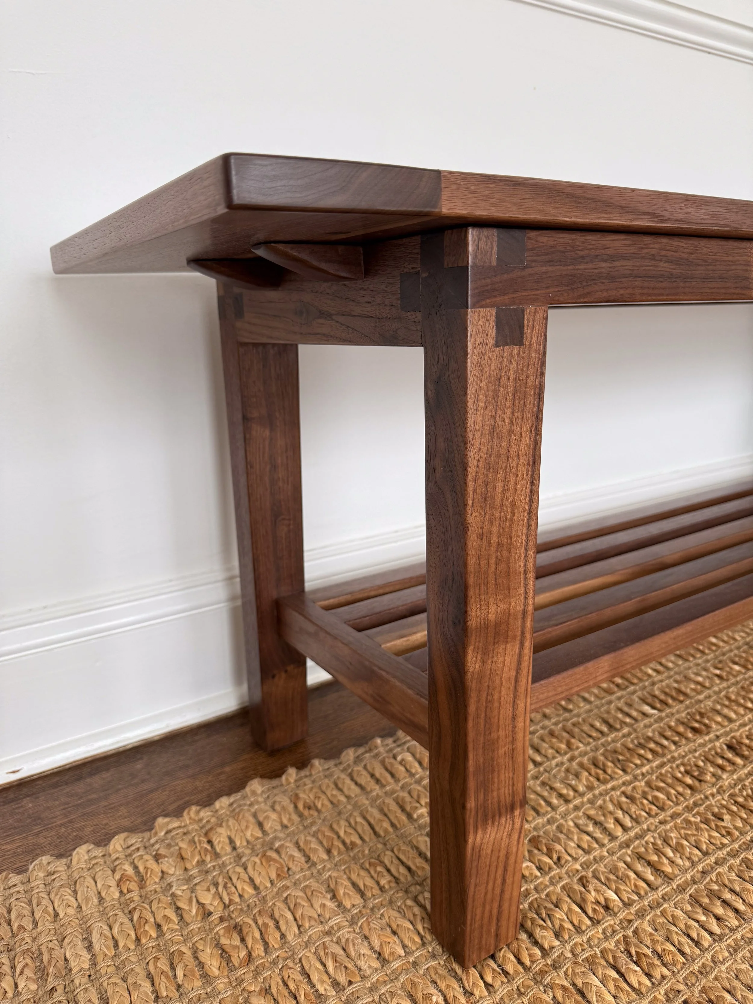 A close-up view of a wooden bench or table with joinery details, situated on a woven rug against a white wall with decorative molding.