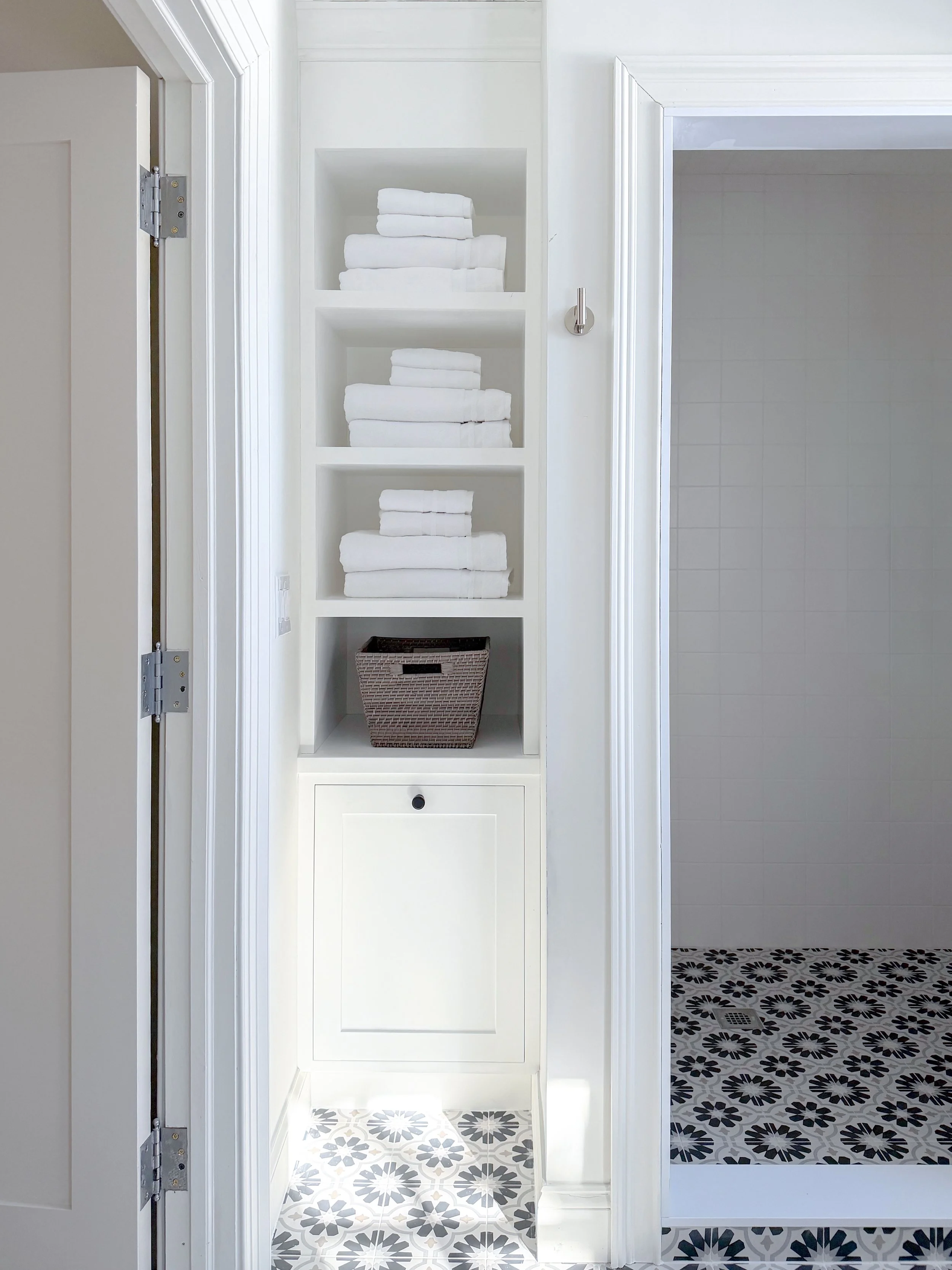 Bathroom with white shelves holding folded white towels, a small basket, and a white cabinet, next to a shower area with patterned black and white floor tiles.
